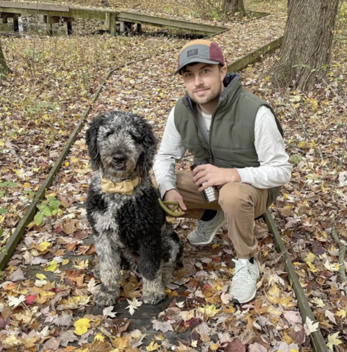A person sitting on a leaf-covered wooden path in a forest next to a curly-haired black and gray dog wearing a yellow neckerchief. The person is wearing a cap, green vest, and holding a coffee cup.