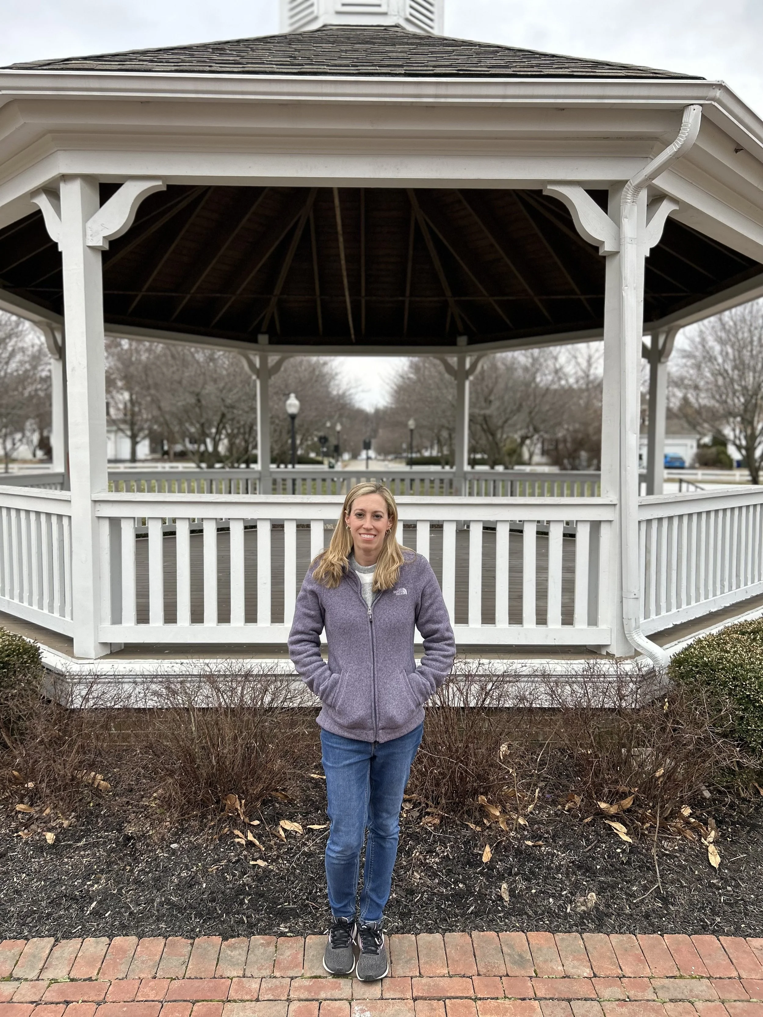 A person in a gray jacket and blue jeans stands in front of a white gazebo on a cloudy day, surrounded by bare bushes and a brick pathway.