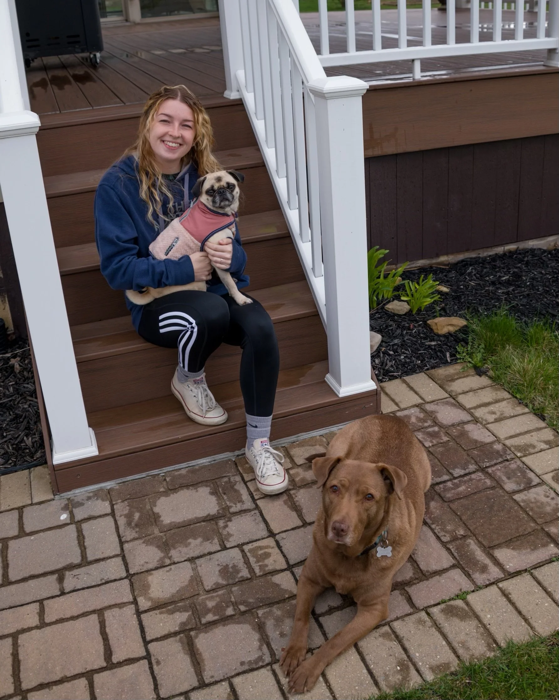 A person sitting on outdoor steps holding a pug wearing a pink jacket, with a brown dog lying on the stone pavement below.