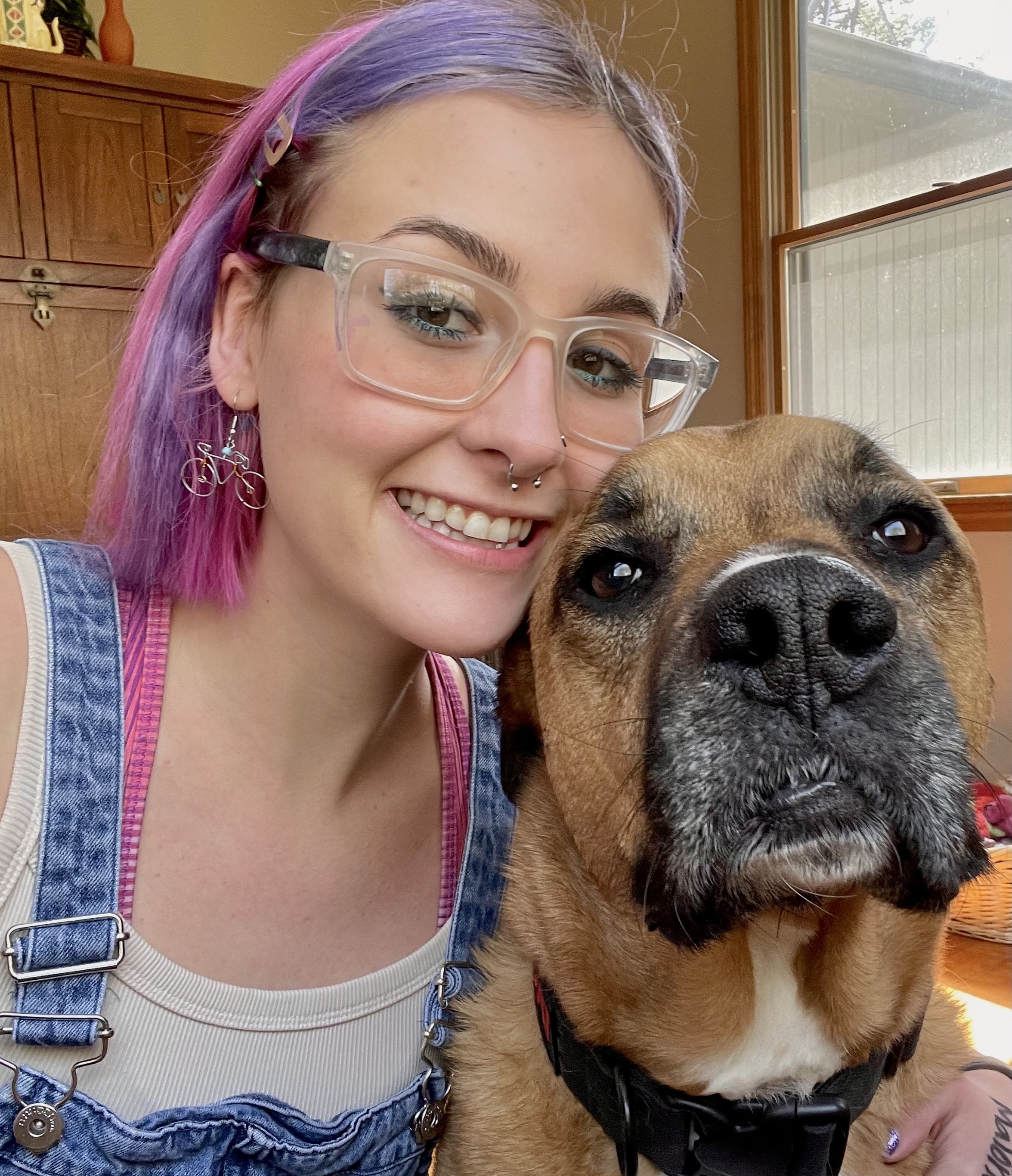 Person with purple hair and glasses smiling next to a brown dog, both in a sunlit room.