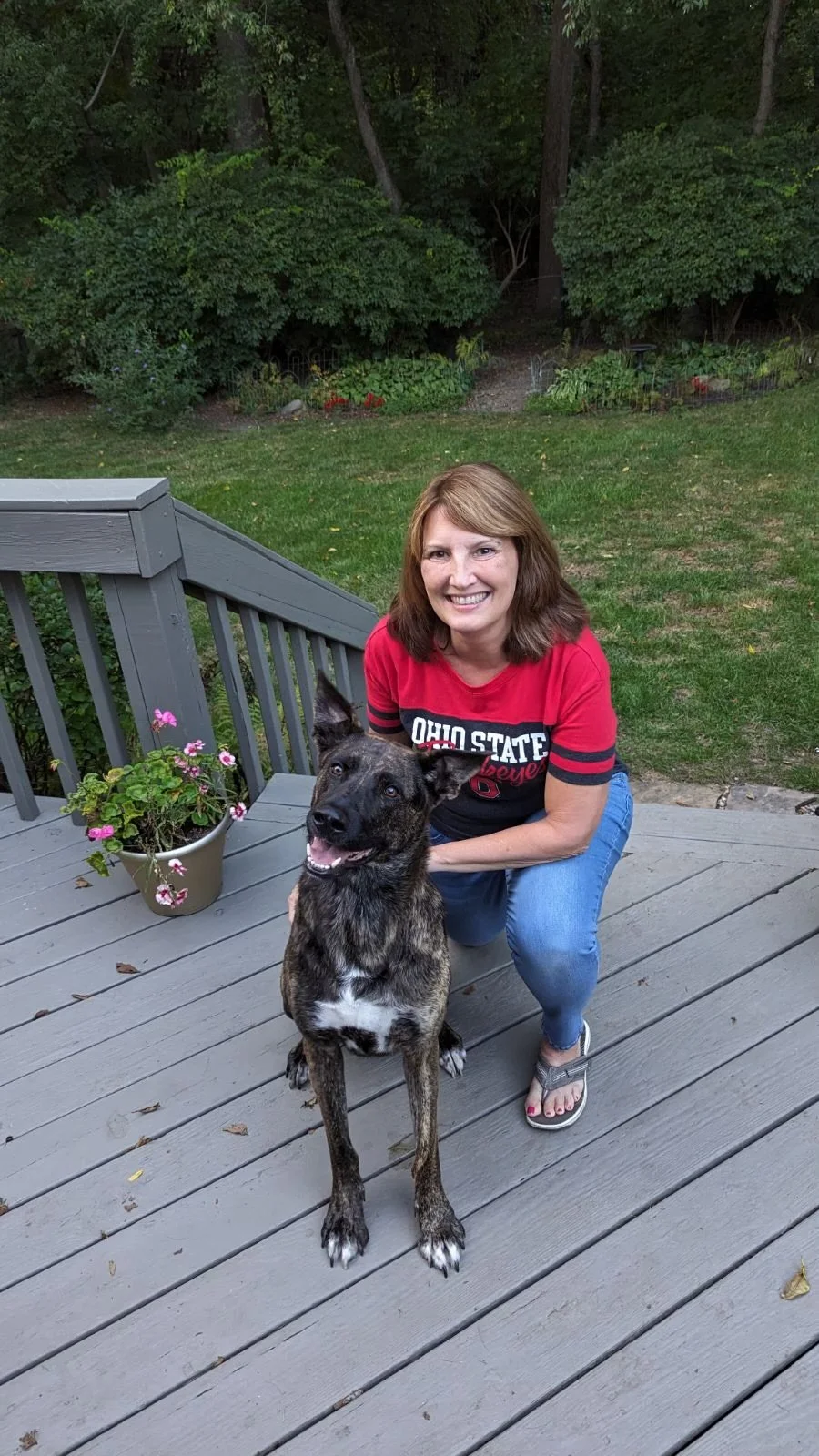 Woman in an Ohio State shirt kneeling on a deck with a German Shepherd mix dog, surrounded by greenery.