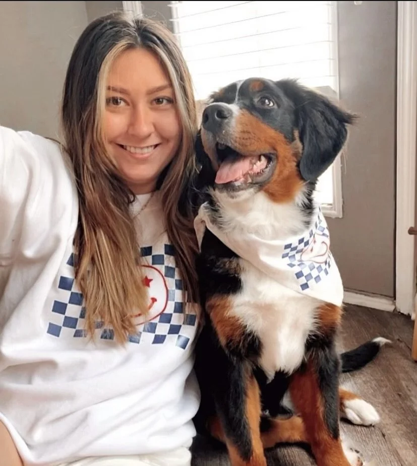 Smiling woman with a dog wearing matching bandana and shirt featuring a smiley face and checkered pattern.