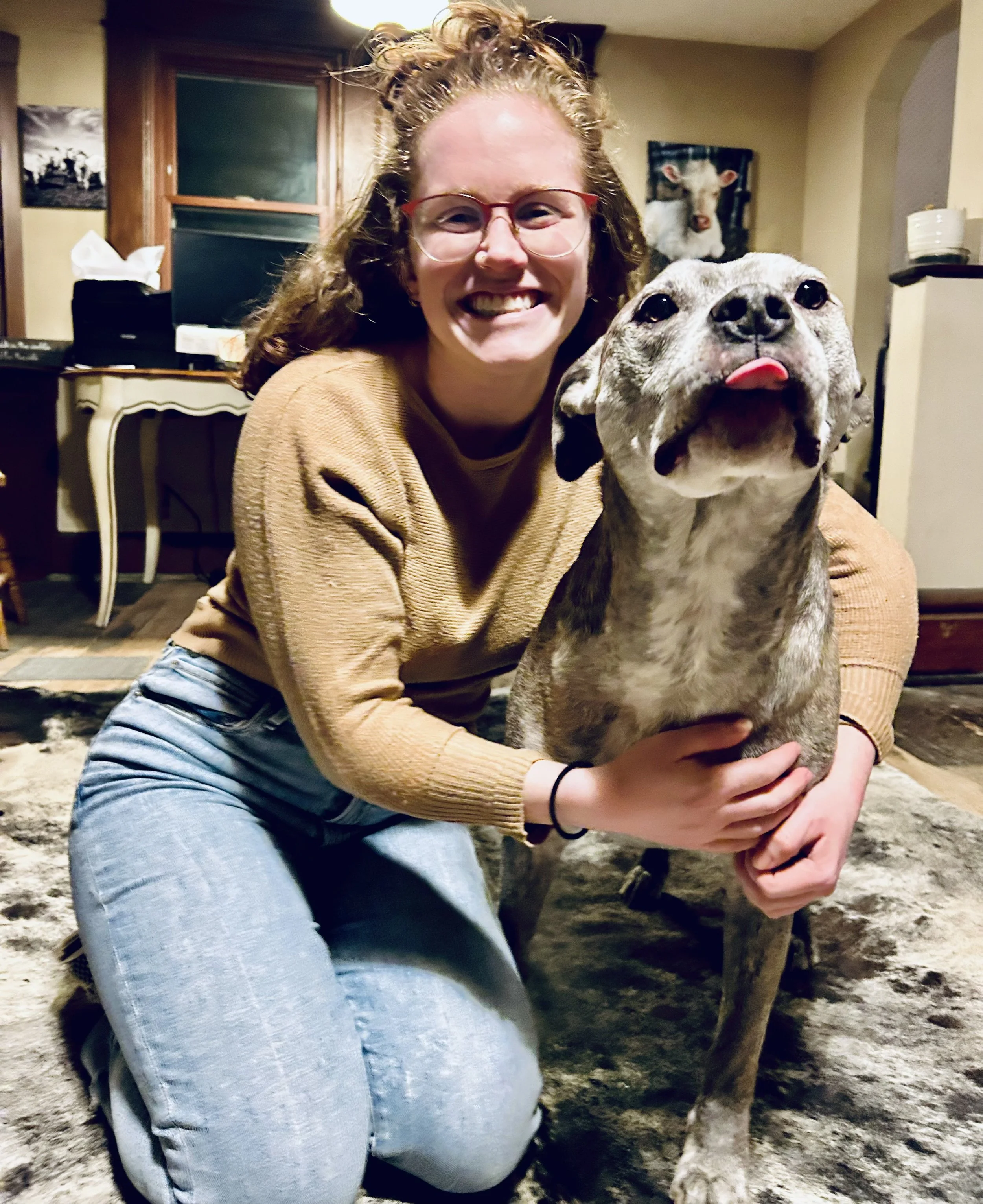 A smiling person kneeling indoors next to a brindle dog with its tongue out.