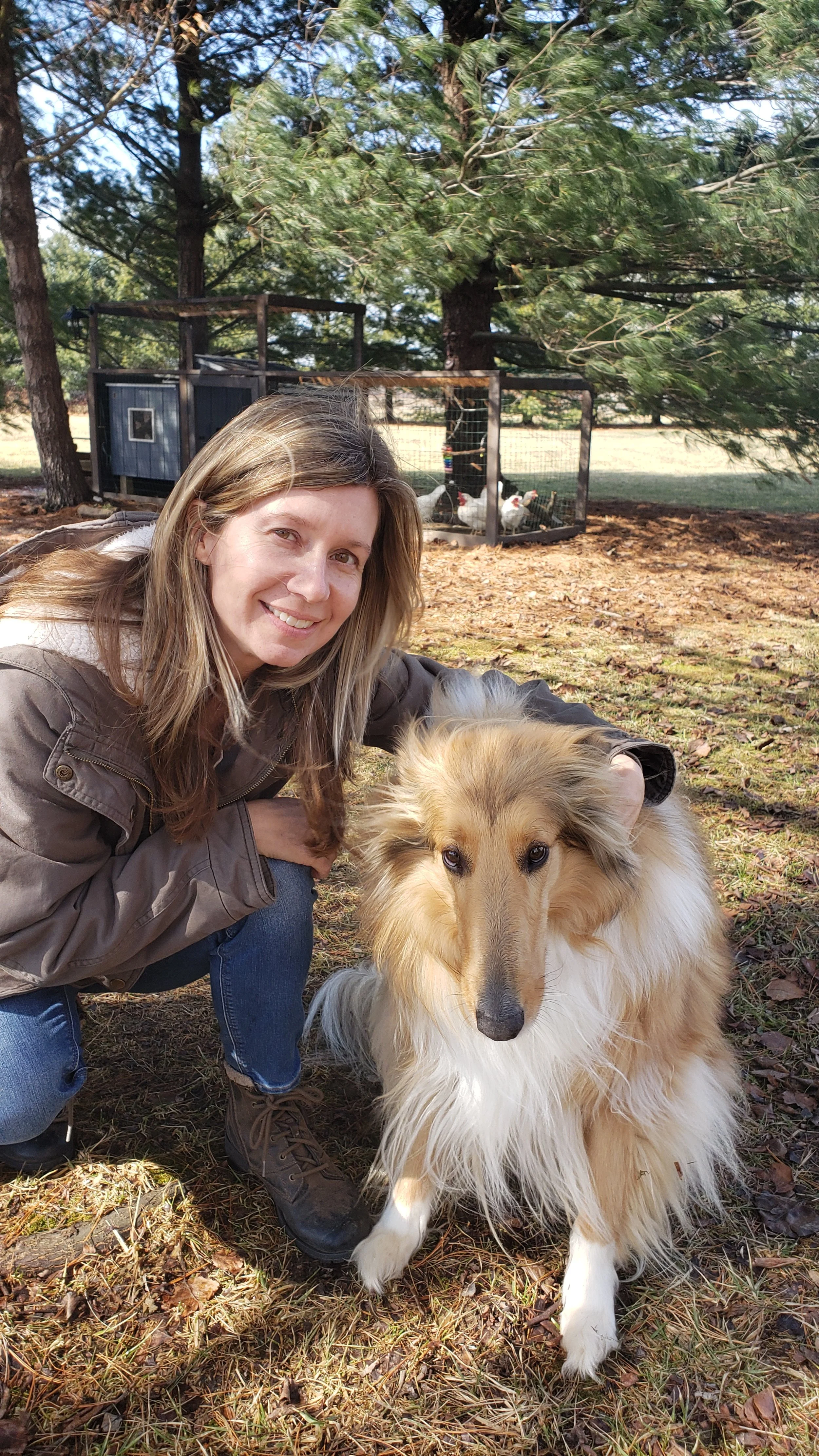 A woman kneeling and smiling next to a rough collie dog in an outdoor setting with trees and a chicken coop in the background.