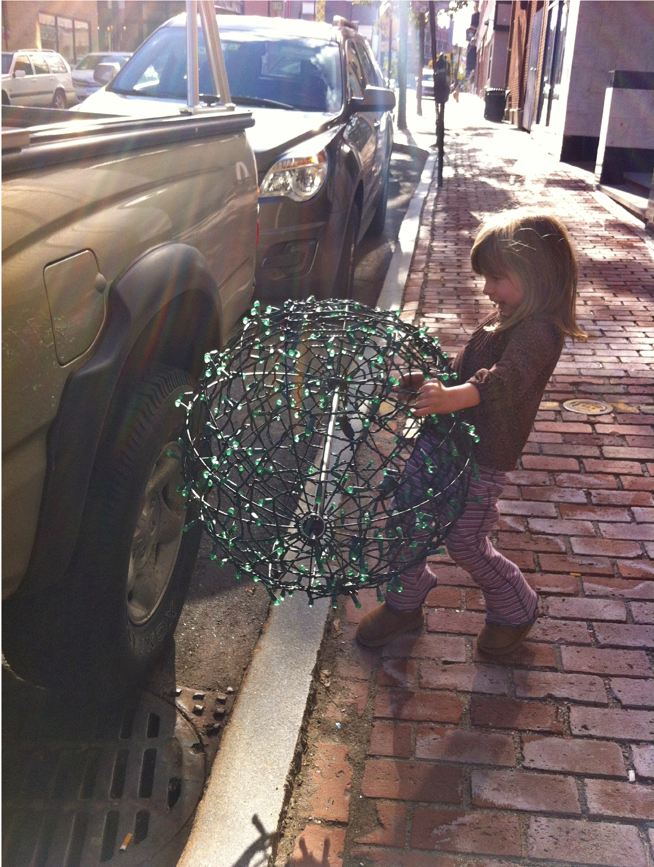 A young girl with light brown hair, wearing a dark sweater, striped pants, and brown boots, is holding a spherical wire frame with small green lights attached, on a brick sidewalk near parked cars on a city street.