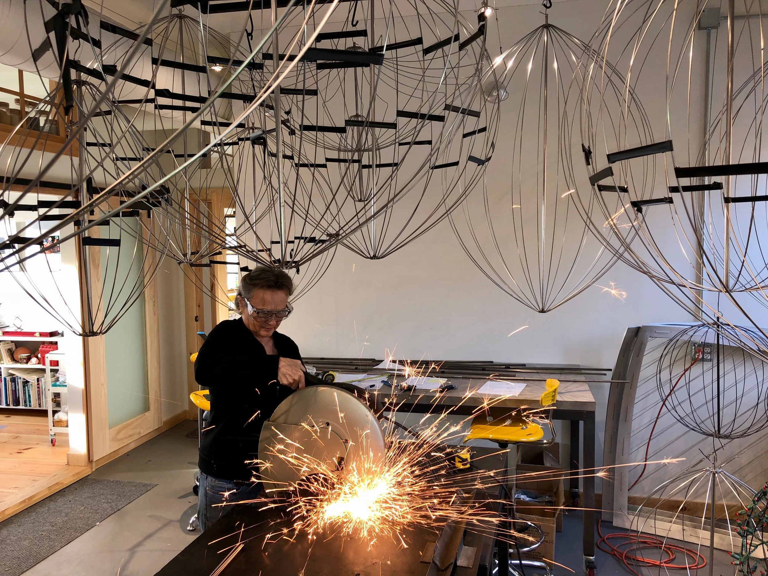 A woman working with sparks flying from a power tool in a workshop or studio, surrounded by suspended wireframe geometric sculptures.