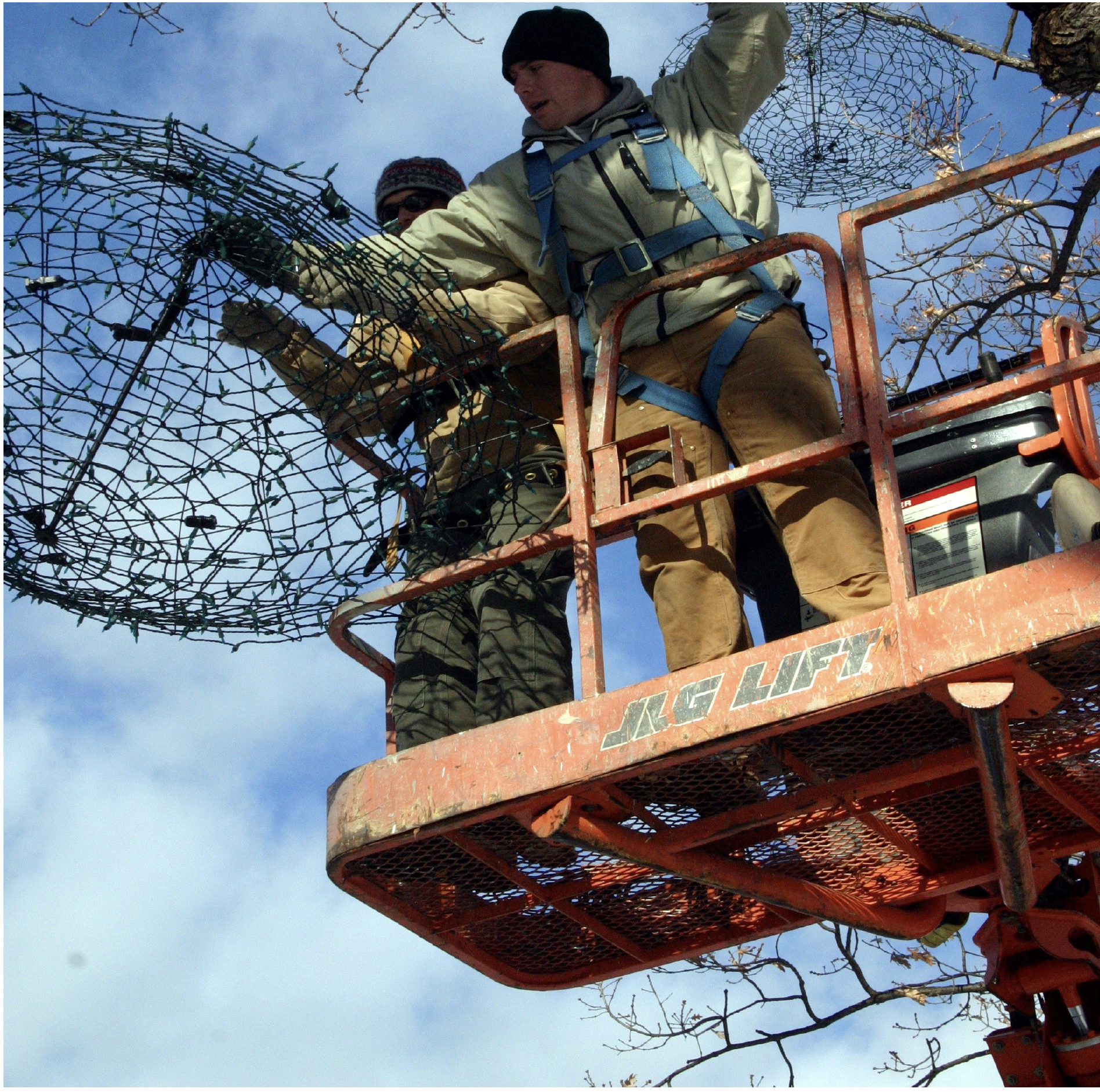Two workers in a cherry picker lift are installing or repairing Christmas lights on a tree on a cloudy day.
