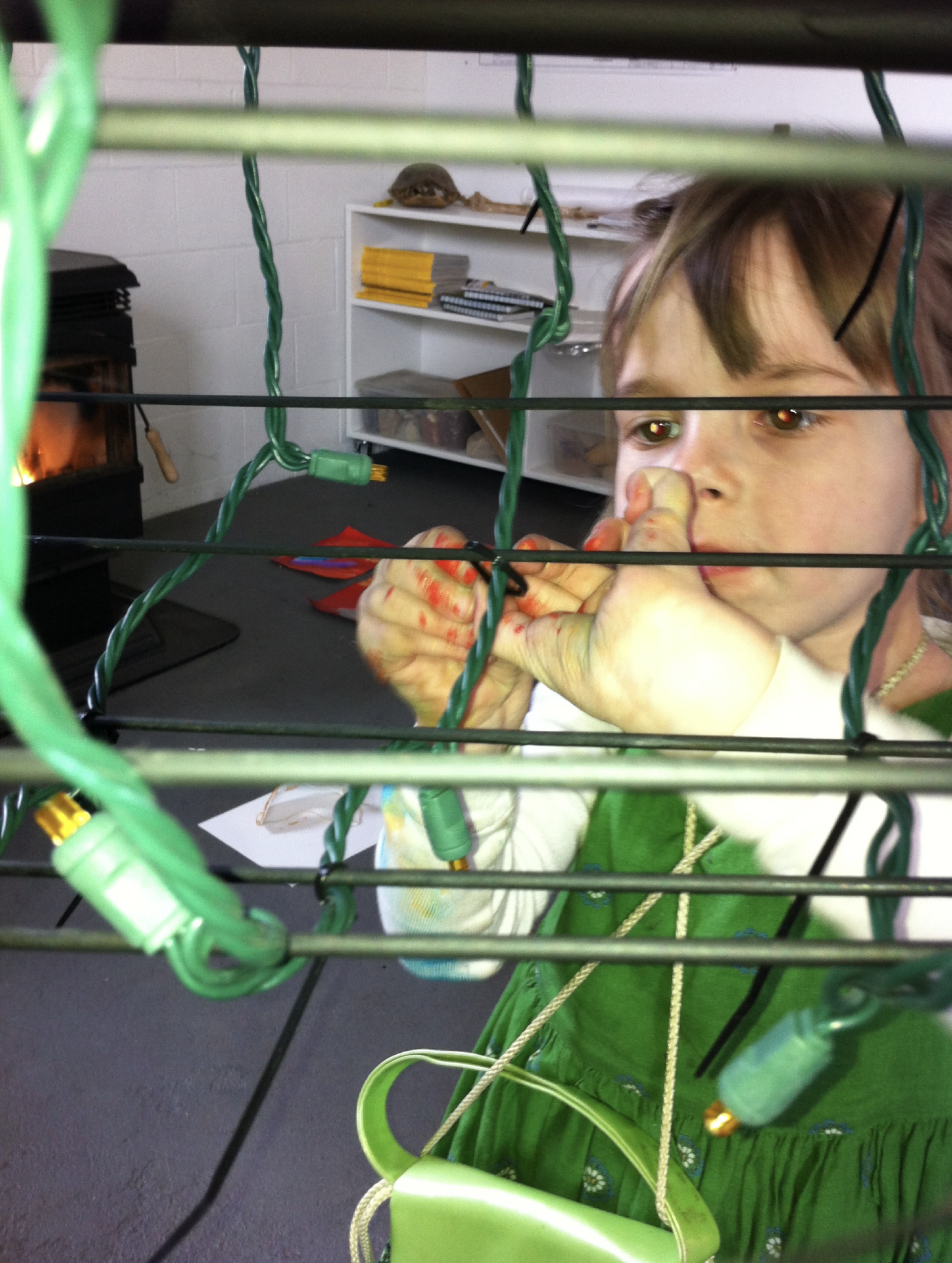 A young girl with colorful hands is looking through a wire fence, possibly at a Christmas tree with lights, in a room that has a white shelf and a small heater in the background.