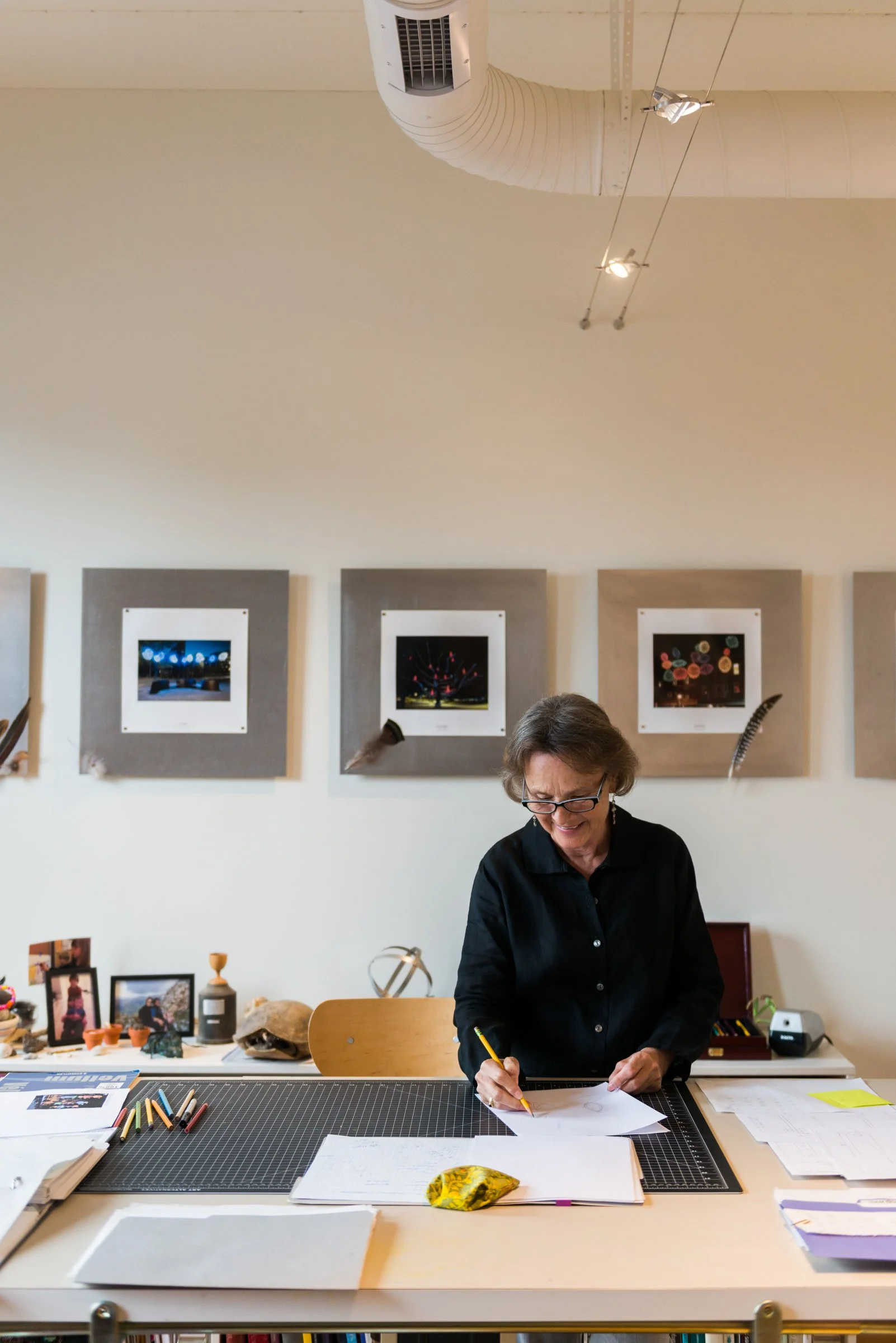 A woman with glasses, wearing a black shirt, is standing at a large desk, writing on paper with a yellow pencil. The desk is filled with papers, notebooks, and colored pencils. Behind her, there are framed photographs and artwork, and a wall decorate