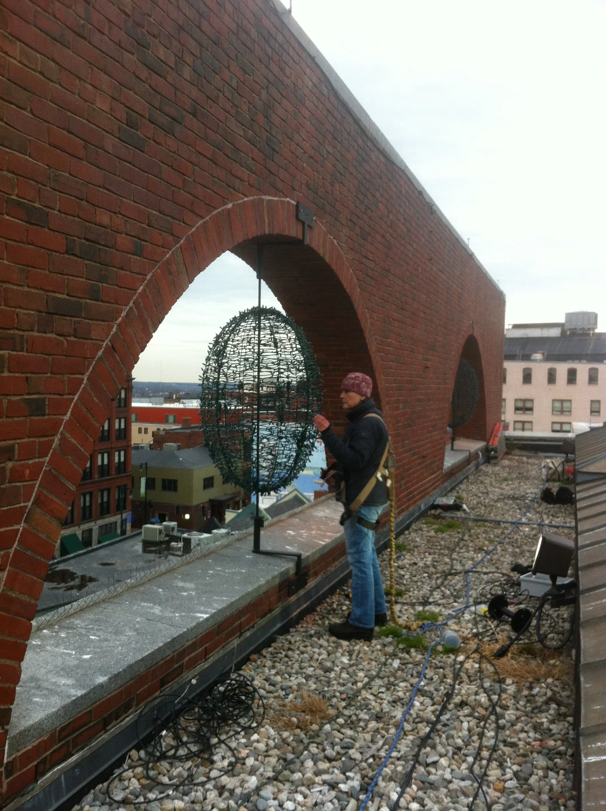 A woman standing on a rooftop next to a brick wall with arches, looking at a decorative lantern.
