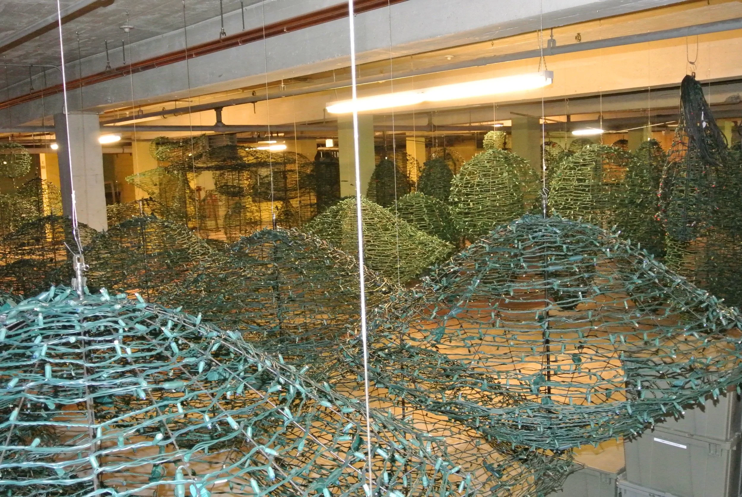 Indoor installation of numerous large, tangled wire sculptures resembling nests or mounds, suspended from the ceiling, with industrial-style lighting and exposed pipes above.