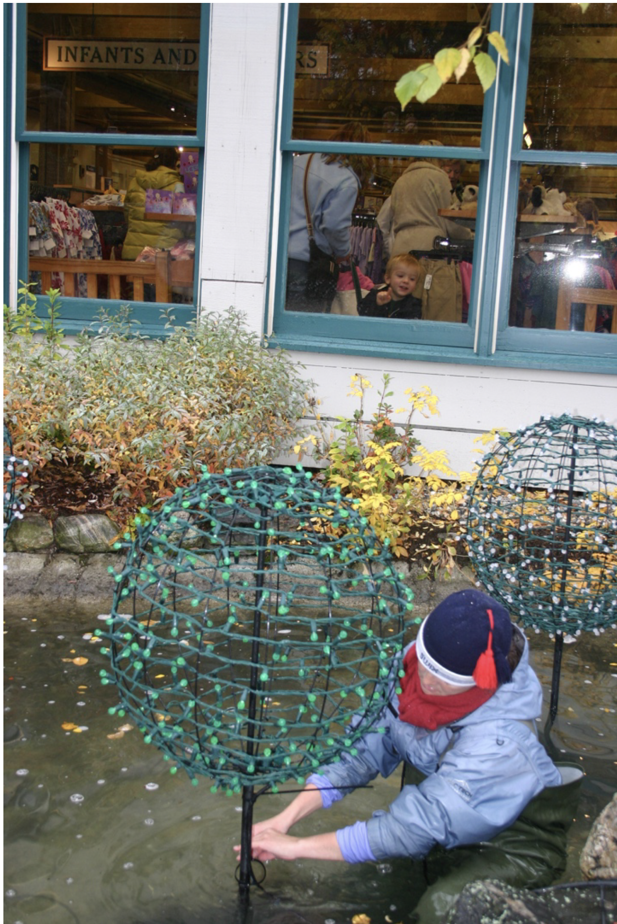 A person in rubber waders and a winter hat working in a small pond, adjusting a large wire-frame sphere with green lights, outside a building with people inside, some of whom are looking out the window at the scene.