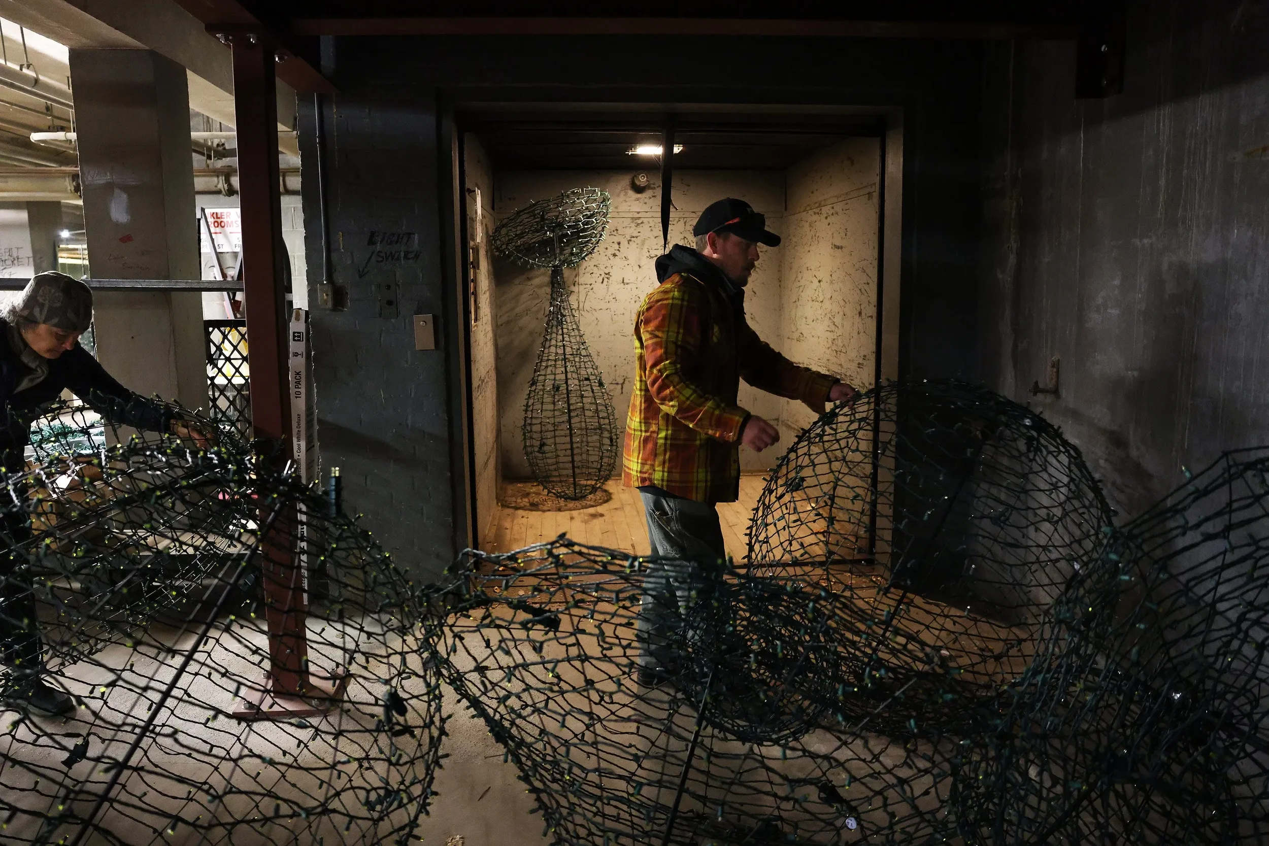 Two people are assembling large wireframe sculpture light displays in a dark indoor storage area.