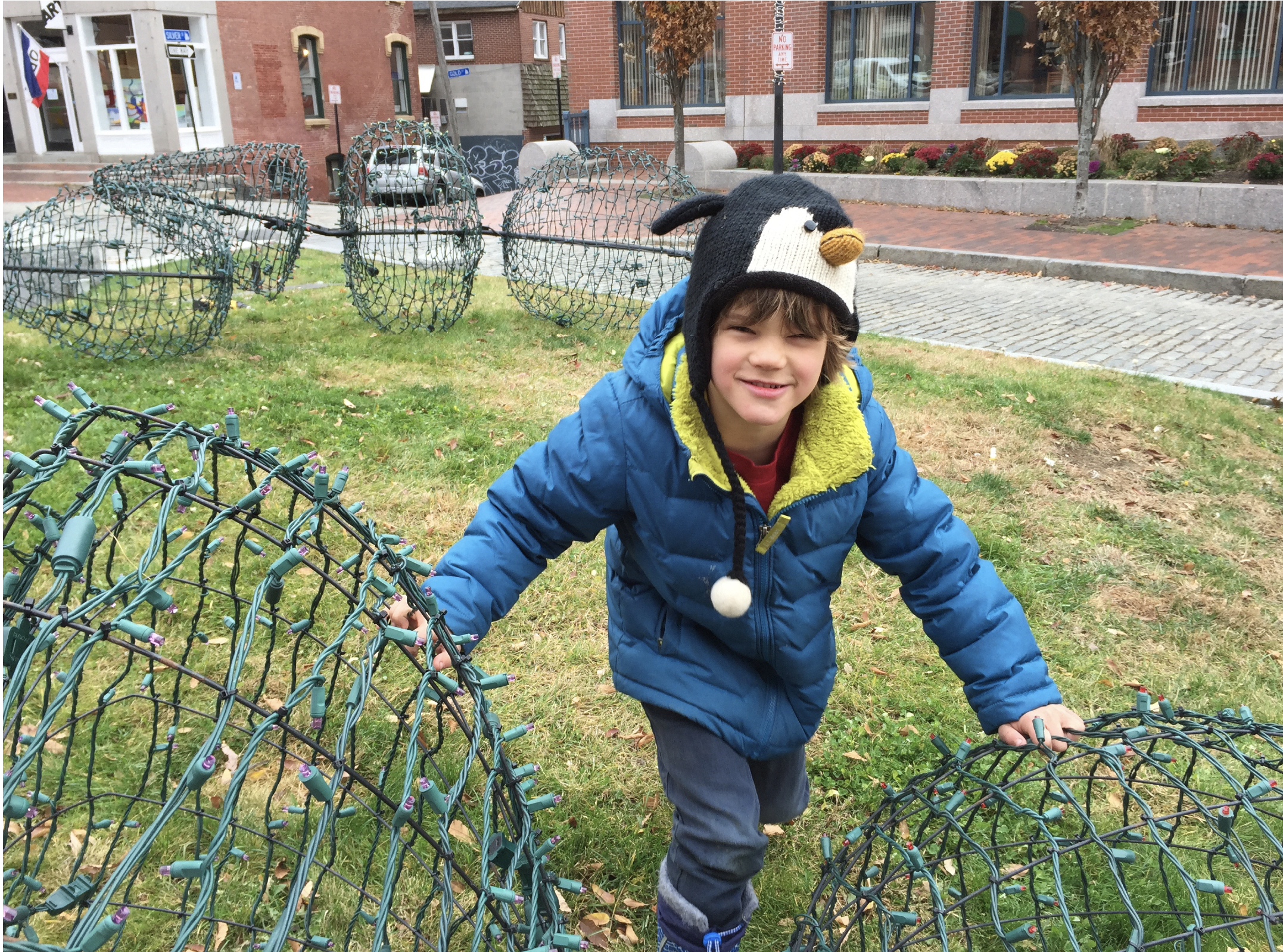 A young boy wearing a penguin hat and a blue jacket, smiling and playing outdoors among large wire-frame holiday light displays on a grassy area in an urban setting.
