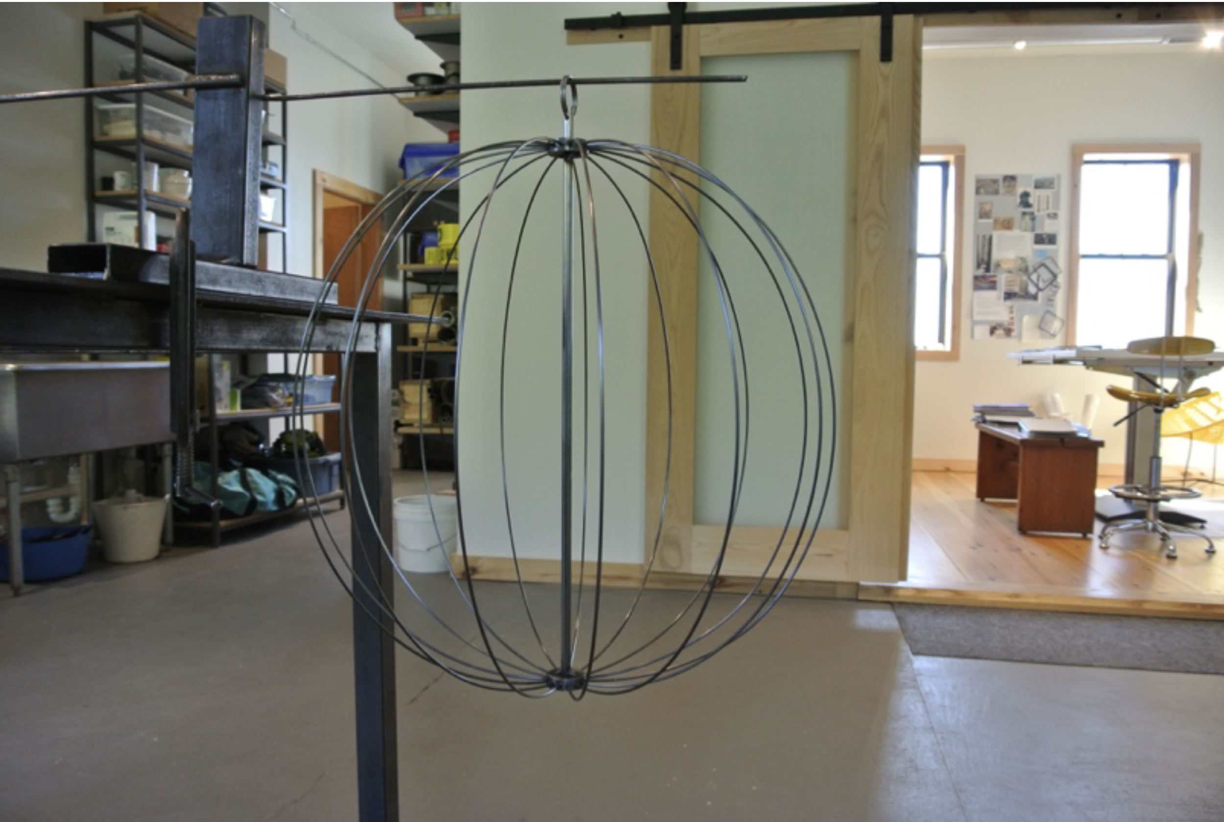 A metal spherical framework, likely a light fixture or art piece, hanging from a ceiling mount in a workshop or studio space.