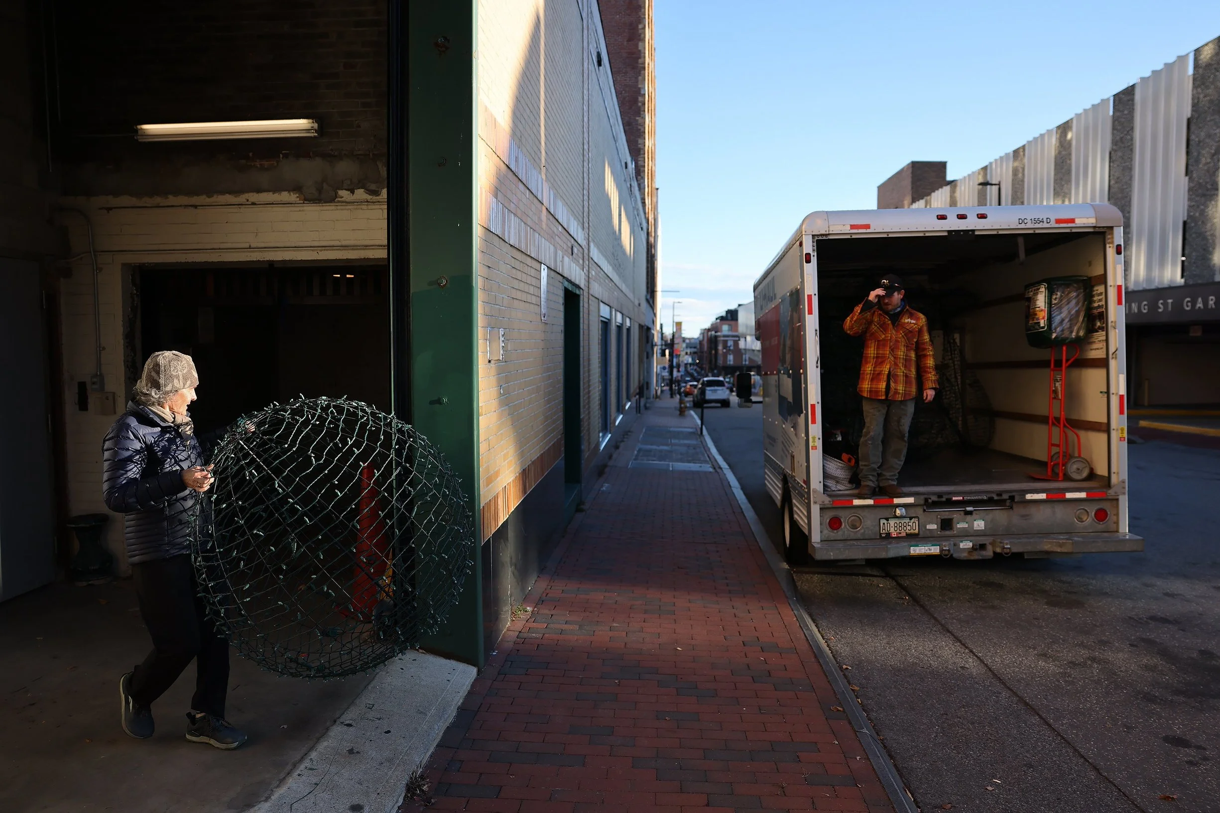 A woman wearing a winter jacket and hat is holding a large wire holiday decoration outside a warehouse. A man in a plaid jacket and cap is standing inside an open delivery truck on the street, which is parked along a brick sidewalk in an urban area.