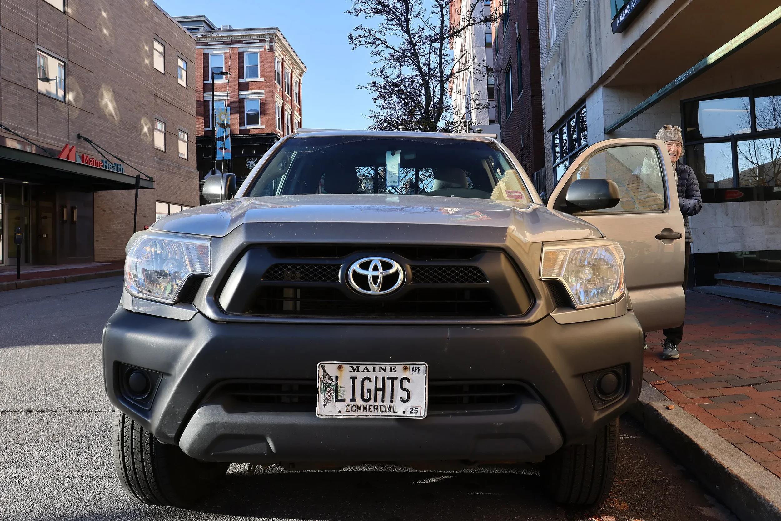 Front view of a black Toyota SUV with a woman standing near the open passenger door on a city street, with buildings and a MaineHealth sign in the background.