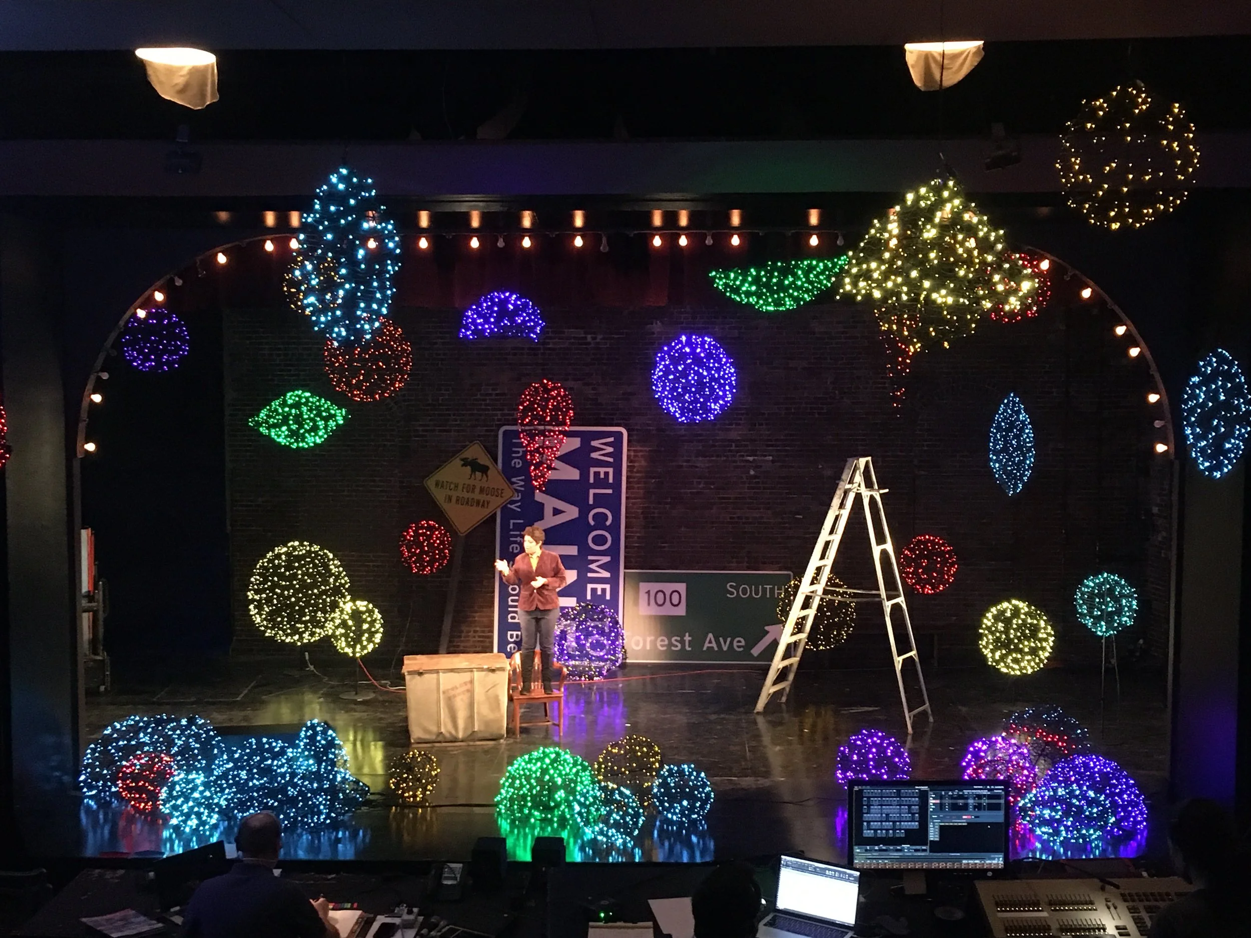 Stage with colorful decorative lights and balloons, ladder, person standing with a microphone, sign reading 'Welcome' and other street signs, and stage equipment.