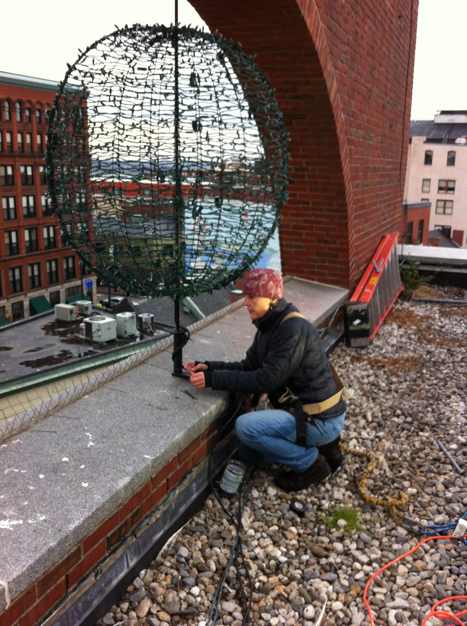 A person wearing a maroon bandana, black jacket, and blue jeans crouches on a gravel rooftop working on a large spherical metal wireframe structure. The rooftop has a brick wall, city buildings, and rooftop equipment in the background.