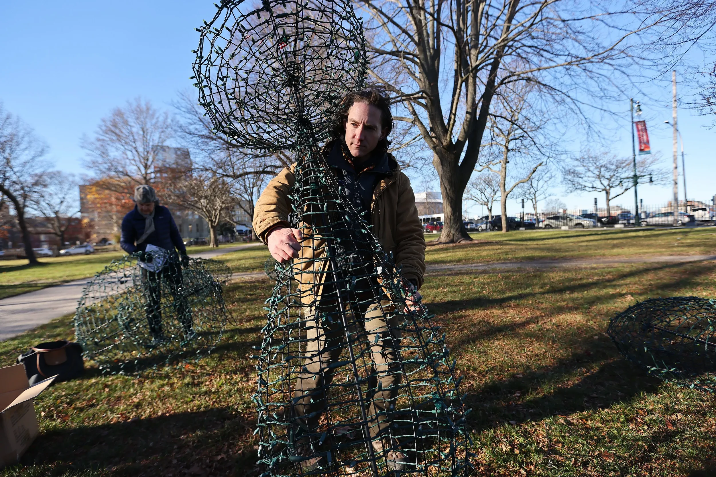 Two people assembling a large wire frame sculpture outdoors on a sunny day, with leafless trees and a park in the background.