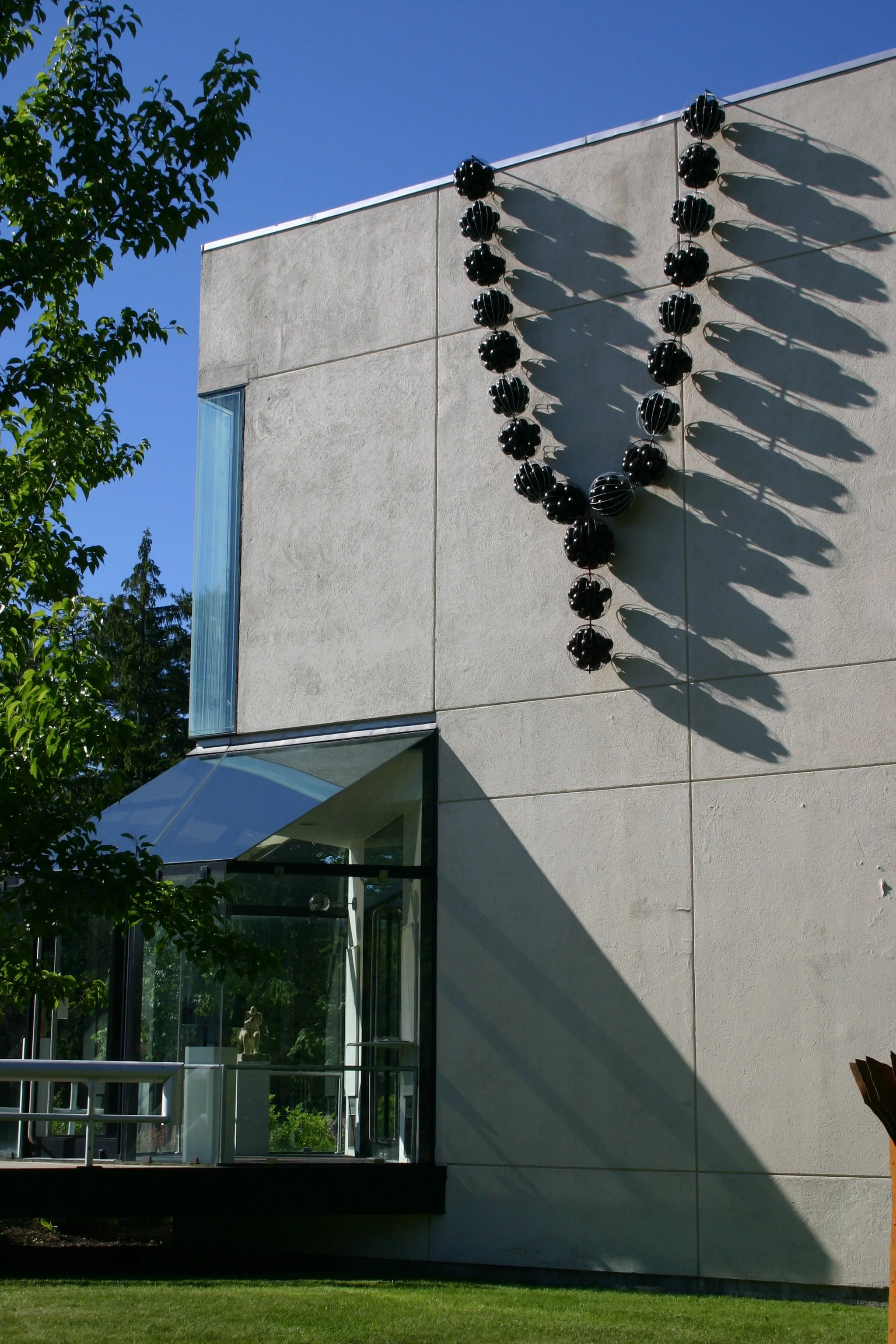 Modern building with beige exterior walls, glass entrance, and black spherical outdoor art sculpture casting shadow on the wall, green grass and trees in the surroundings under a clear blue sky.