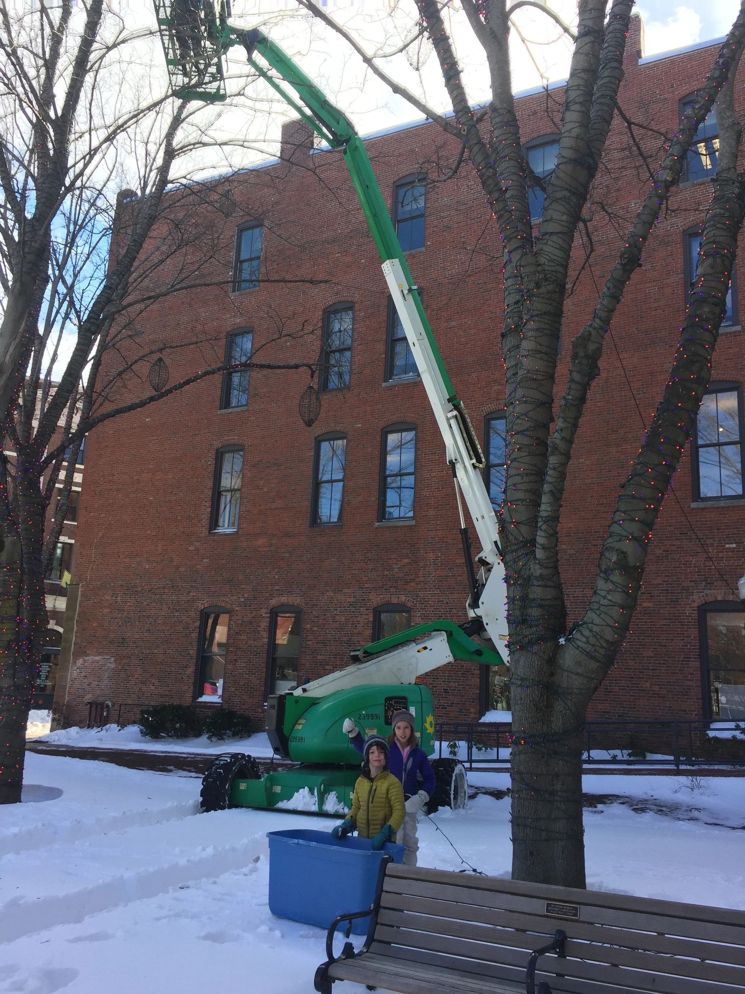 Two children stand in the snow next to a large, green cherry picker lift, which is extended to a tall tree decorated with Christmas lights, in front of a brick building.