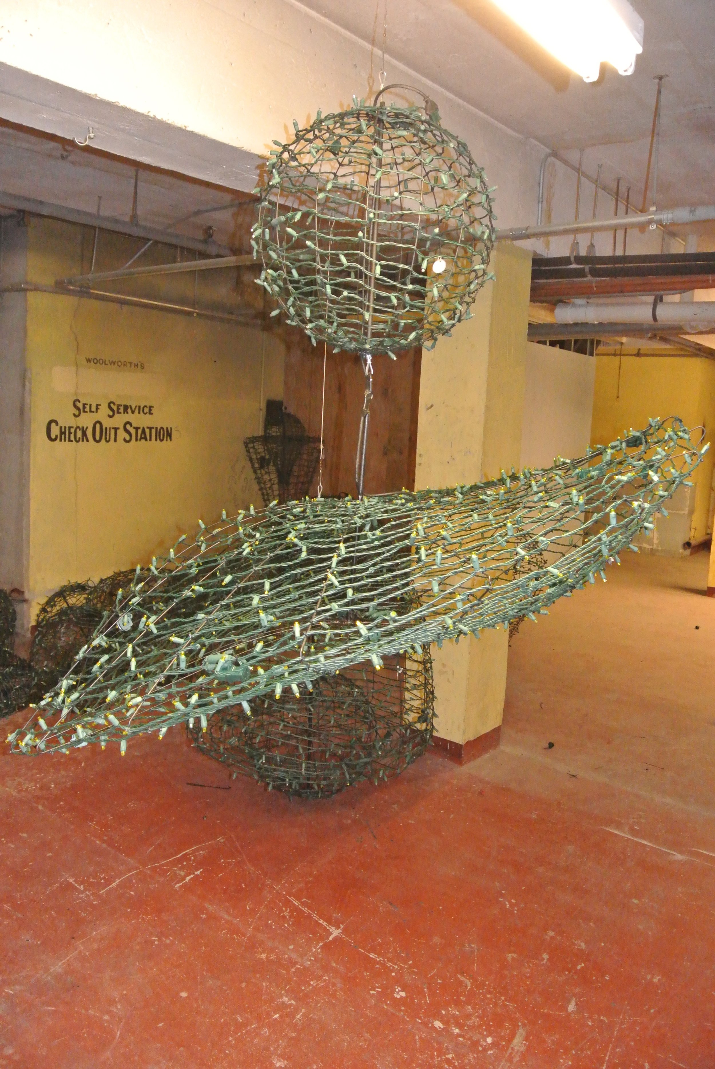 Three large green metal wire sculptures hanging in an industrial-looking room with a yellow wall and a sign that reads 'Self Service Check Out Station'.