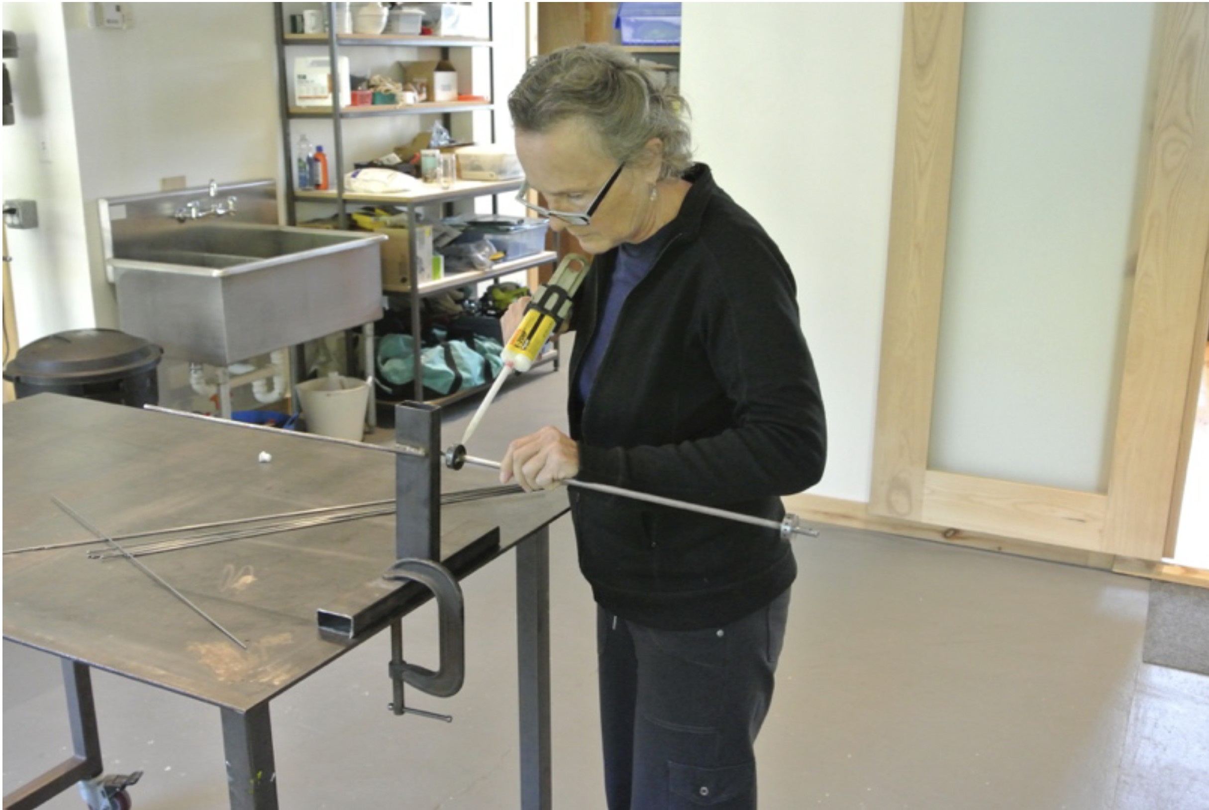 Woman working on a metalwork project in a workshop, using a power tool on a metal rod secured with a clamp.