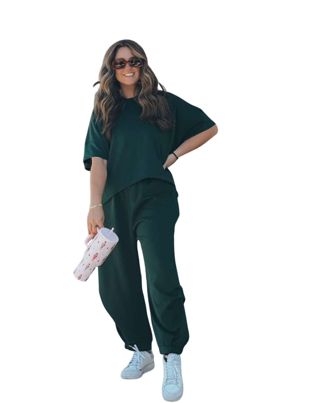 A woman with long wavy hair wearing sunglasses, a loose dark green top, matching green pants, white sneakers, and holding a decorated tumbler, standing with one hand on her hip and smiling.