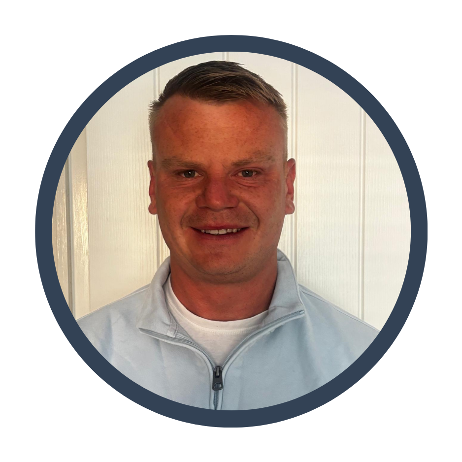 A smiling man with short light brown hair, wearing a white zip-up jacket, standing in front of a white wall.