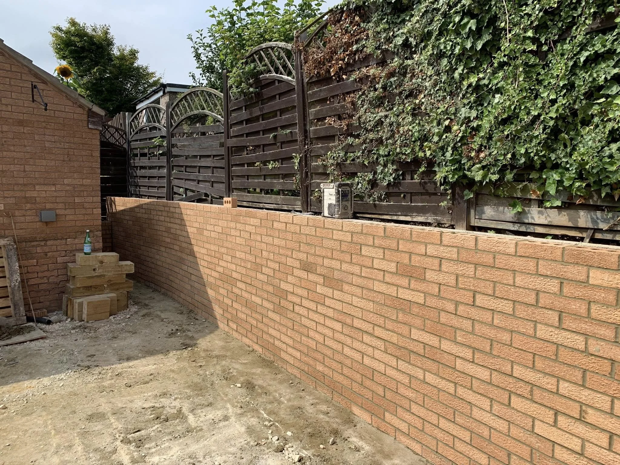 A newly built brick wall with a wooden fence on top, adjacent to a small brick structure in a backyard, with some construction materials and a bottle placed on a corner of the wall, and trees in the background.