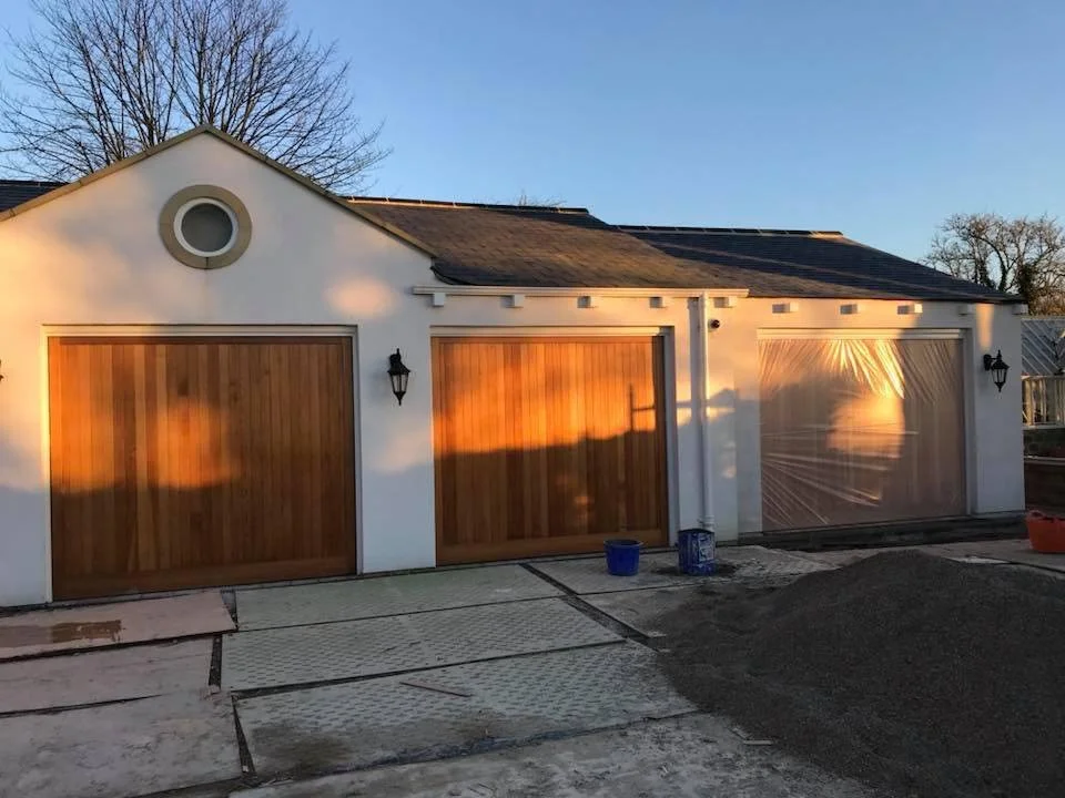 A house with three garage doors, two made of wood and one covered with plastic, during sunset with sunlight reflections on the doors, and construction materials and gravel in front.