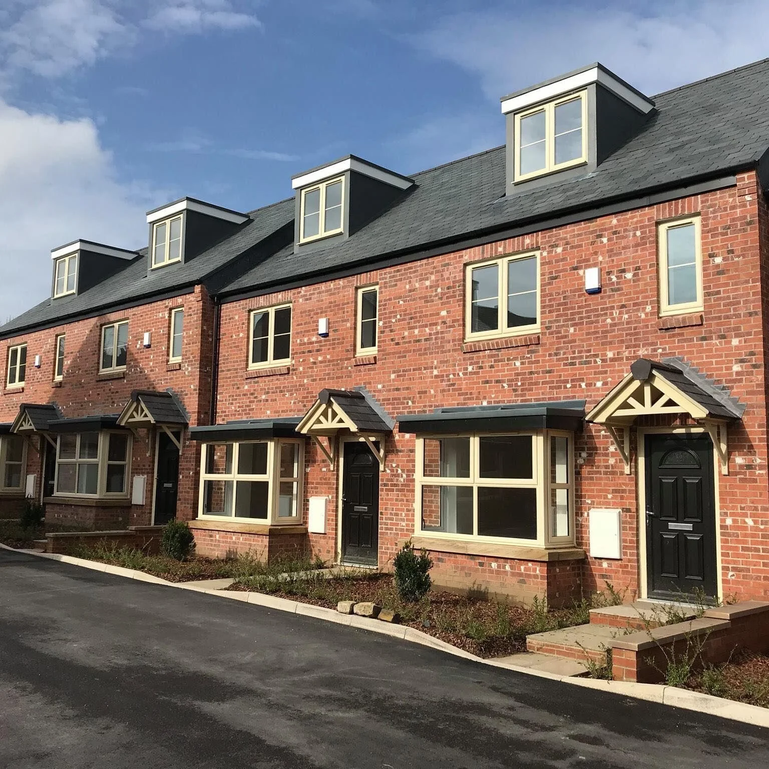 A row of modern red brick townhouses with black doors, bay windows, and dormer windows on the roof, under a blue sky.