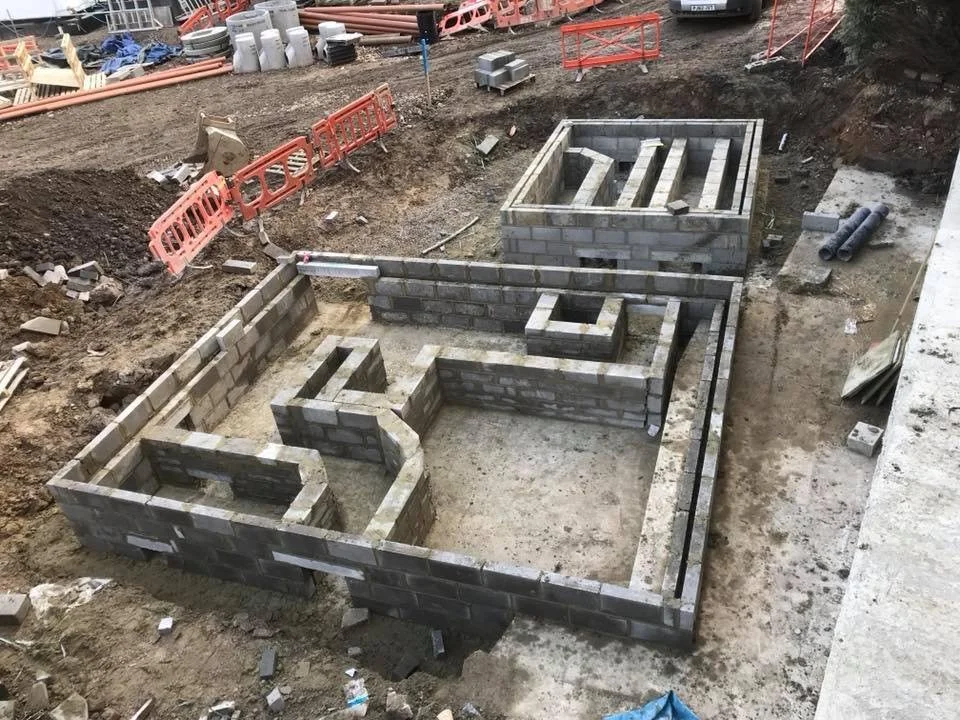 Under construction site showing a foundation with brick walls in progress, with trenches and wooden supports for interior walls, surrounded by construction materials and orange safety barriers.