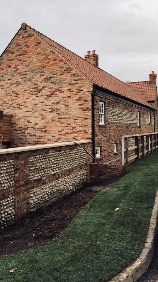 A brick house with a red-tiled roof and small multiple windows, near a brick and pebble wall with a well-maintained grassy lawn in the foreground.