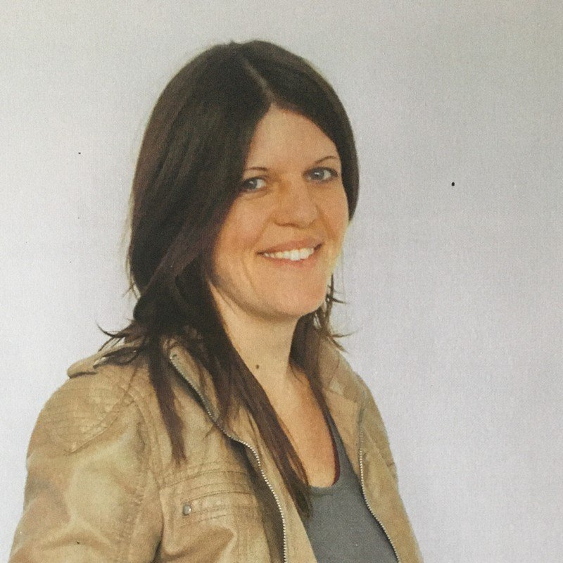 Ein lächelnder Frau mit braunen Haaren und dunklem Pony, trägt eine beige Jacke und ein graues Top, steht vor einem weißen Hintergrund.