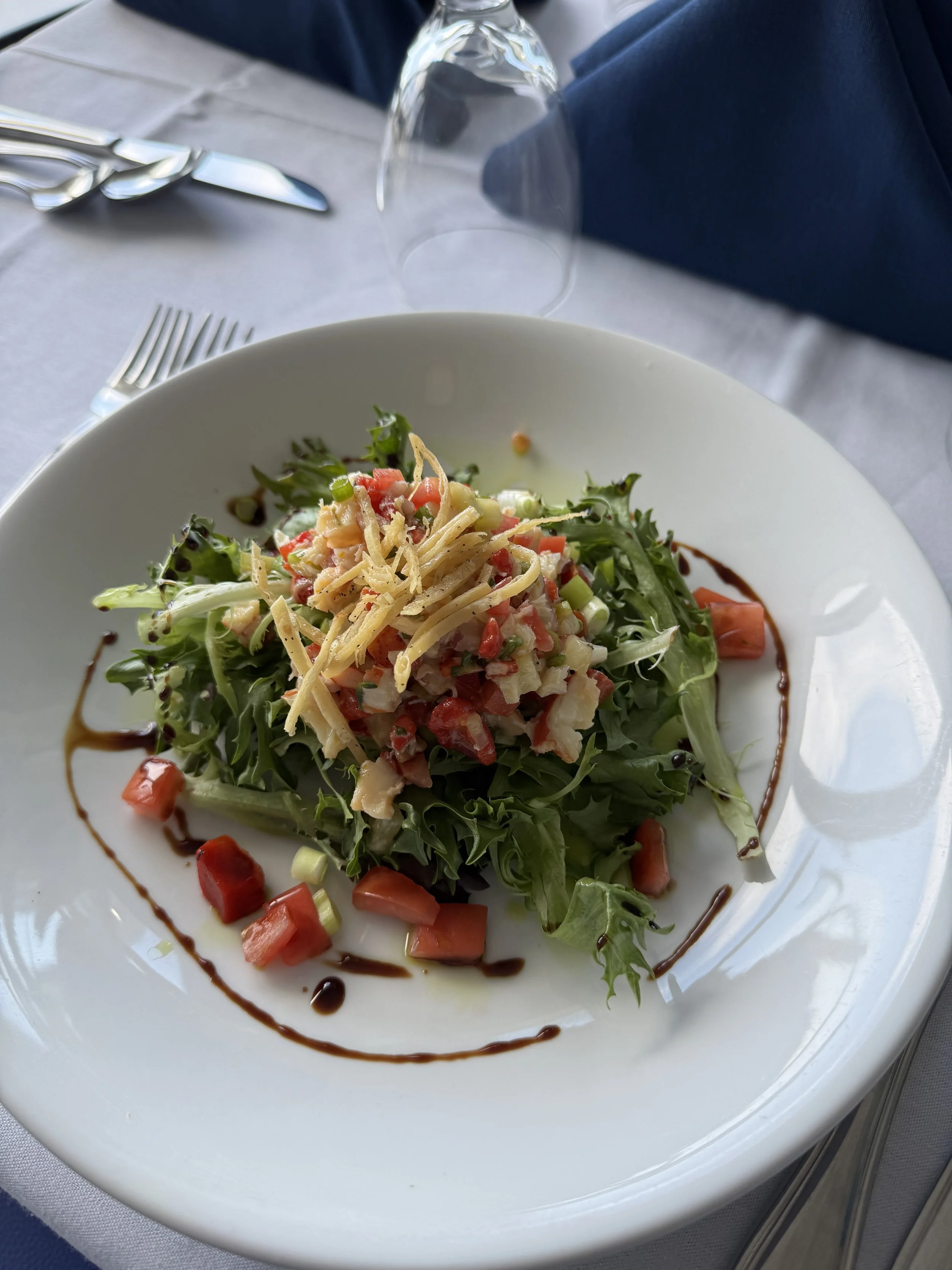 A salad with mixed greens topped with diced tomatoes, shredded cheese, and other vegetables, garnished with a drizzle of balsamic vinegar or dressing, served on a white plate at a restaurant table.