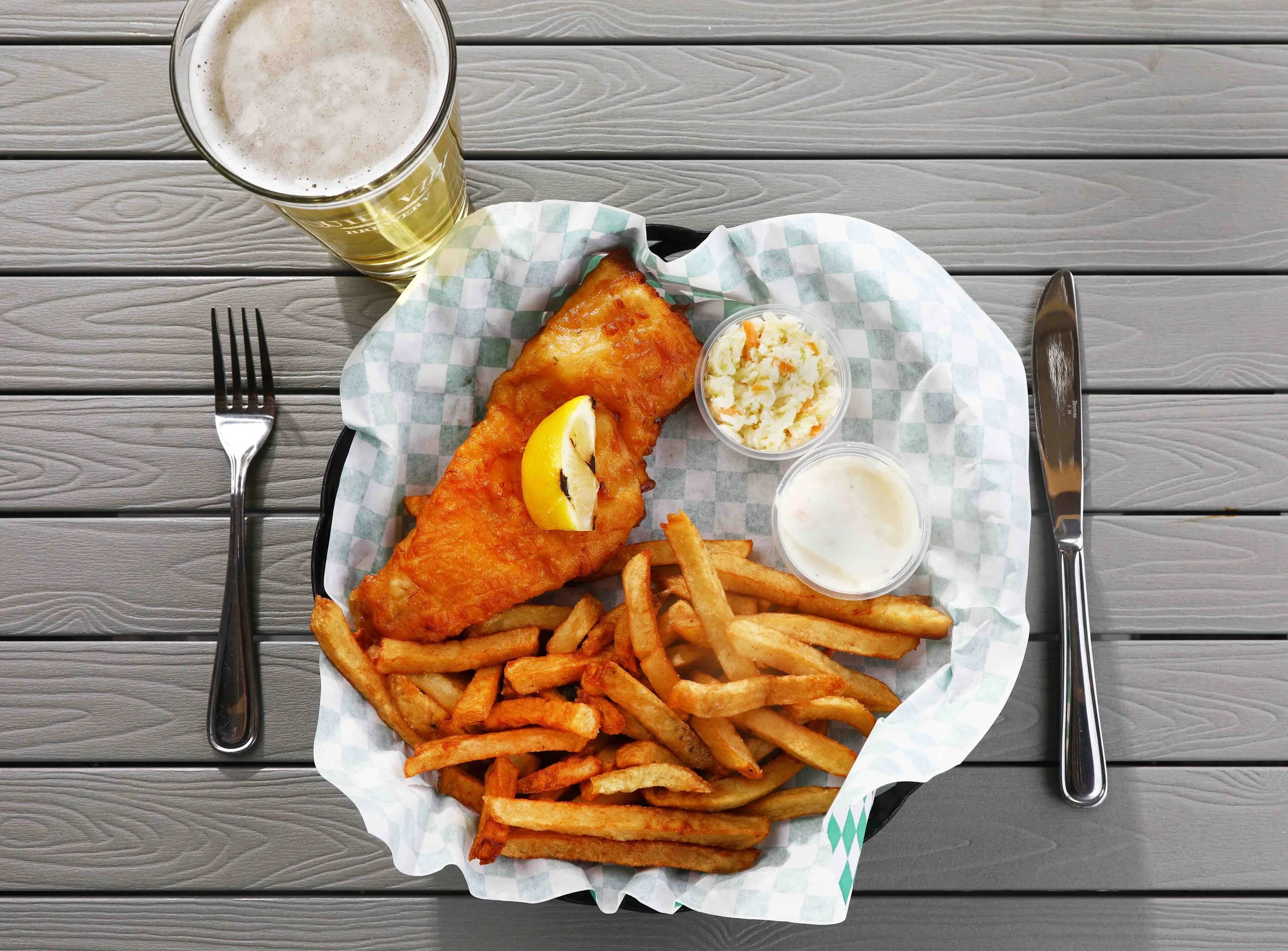 Plate of fried fish with lemon wedge, French fries, two small cups of coleslaw and tartar sauce, glass of beer on gray wooden table.
