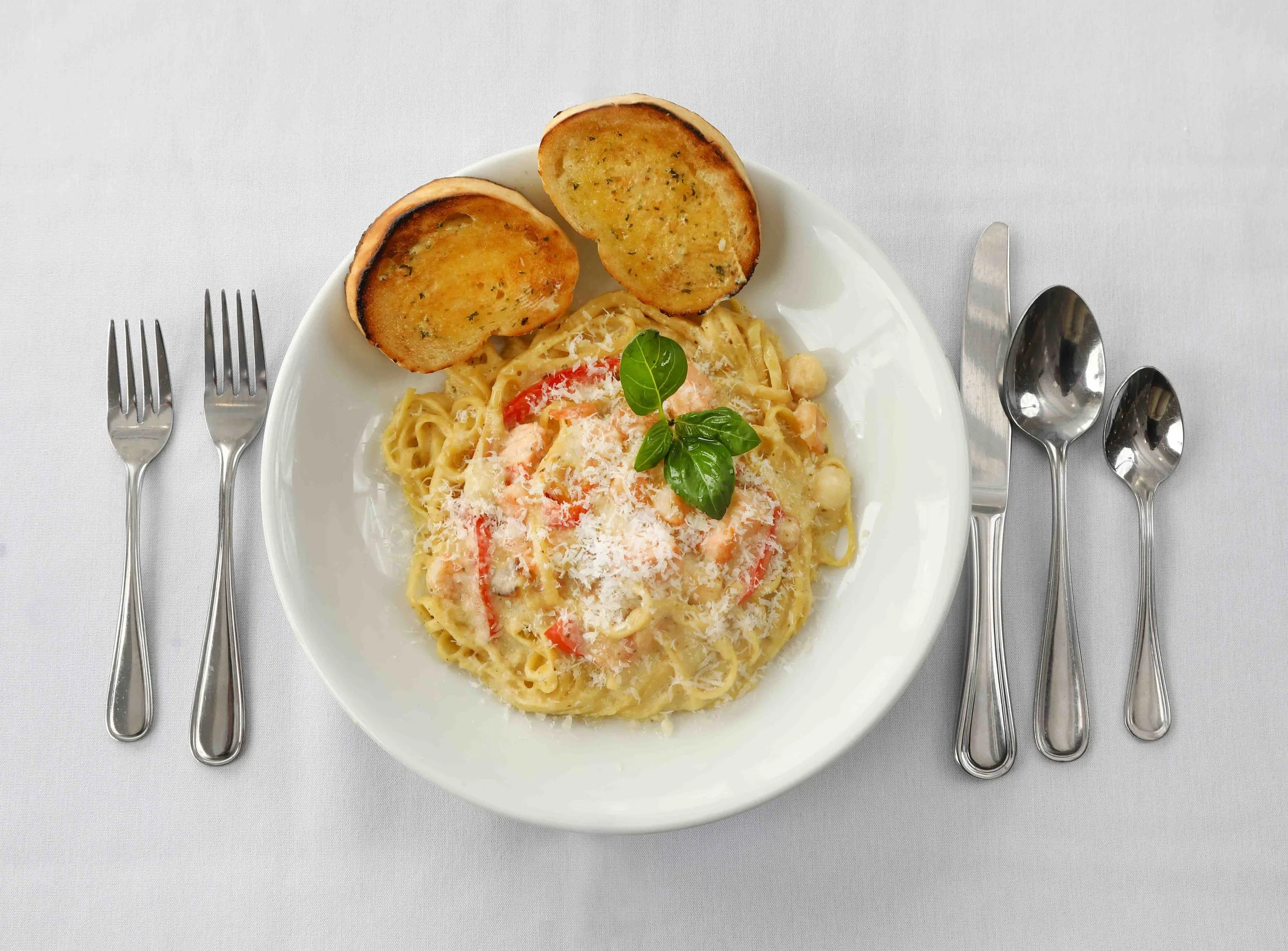 Plate of creamy pasta with red peppers and grated cheese, garnished with basil, served with toasted garlic bread on the side, placed on a white tablecloth with cutlery.
