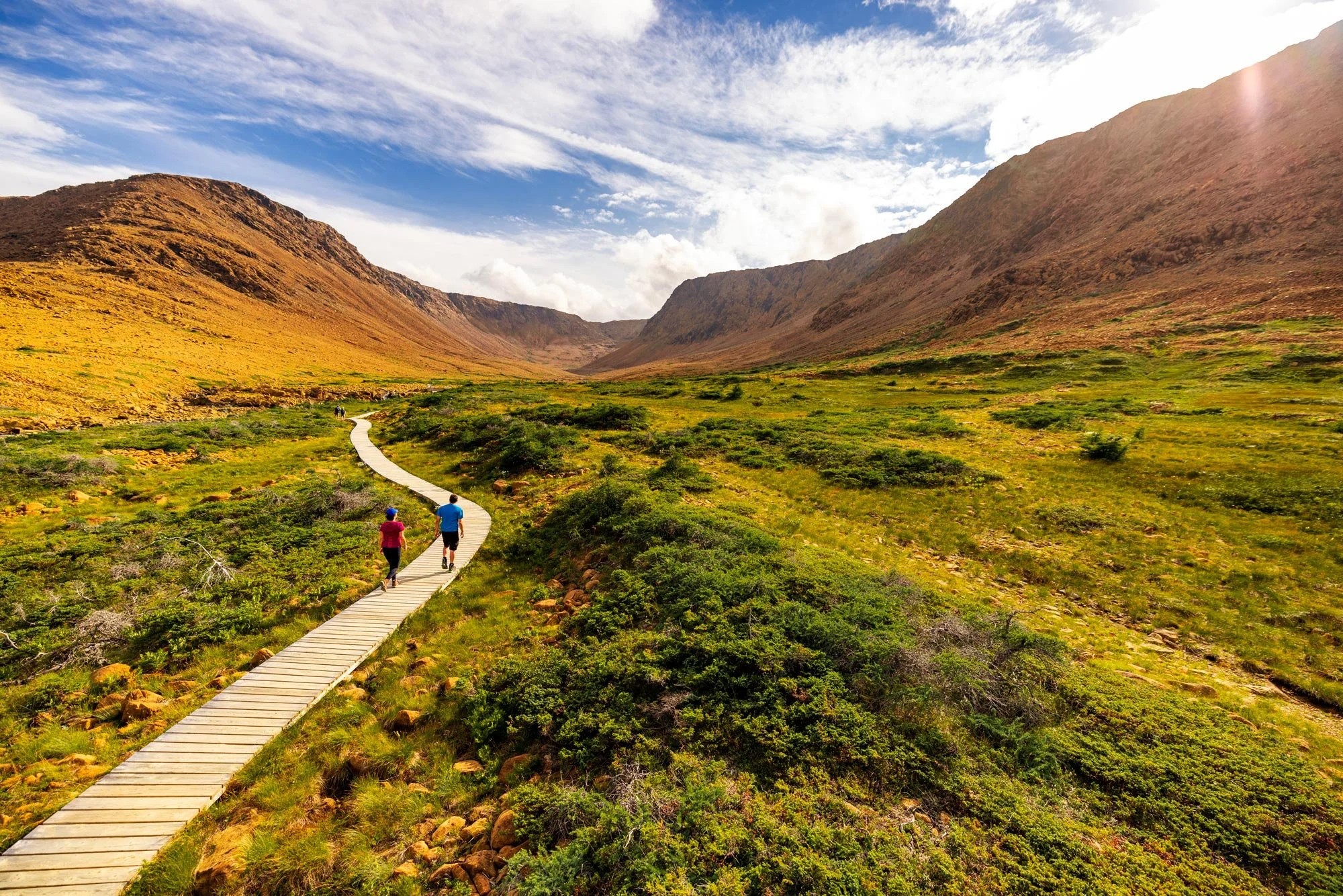 A winding wooden pathway through a lush green valley surrounded by mountains under a partly cloudy sky, with several people walking along the path.