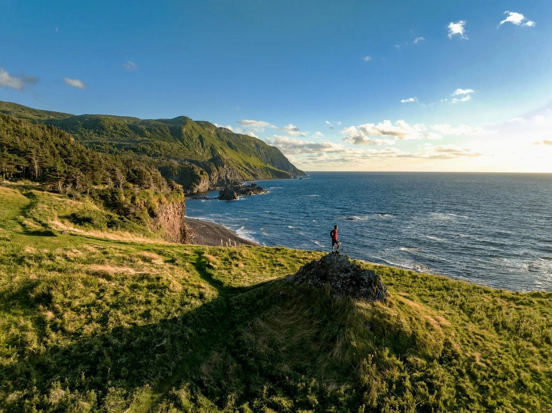 A person standing on a rock overlooking a shoreline with green hills and mountains, ocean, and sky with clouds at sunset.
