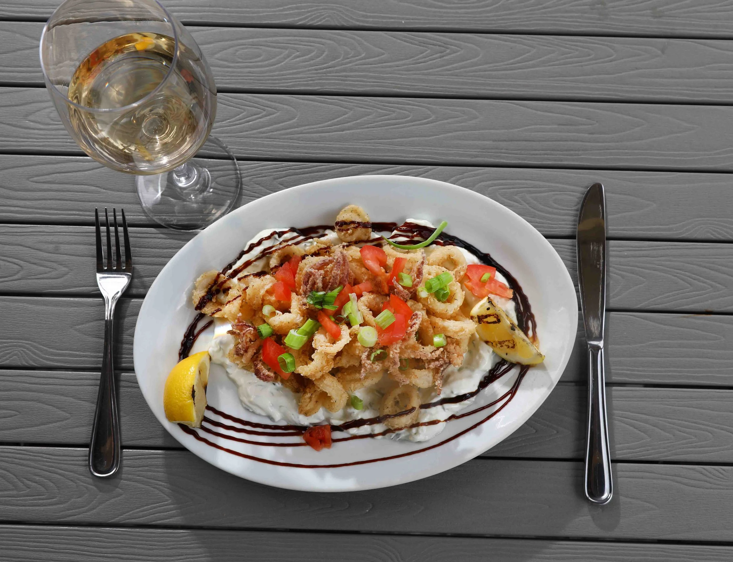 Plate of fried calamari garnished with chopped tomatoes, green onions, lemon wedges, and drizzled with dark sauce, placed on a wooden table with a glass of white wine, fork on the left, and knife on the right.