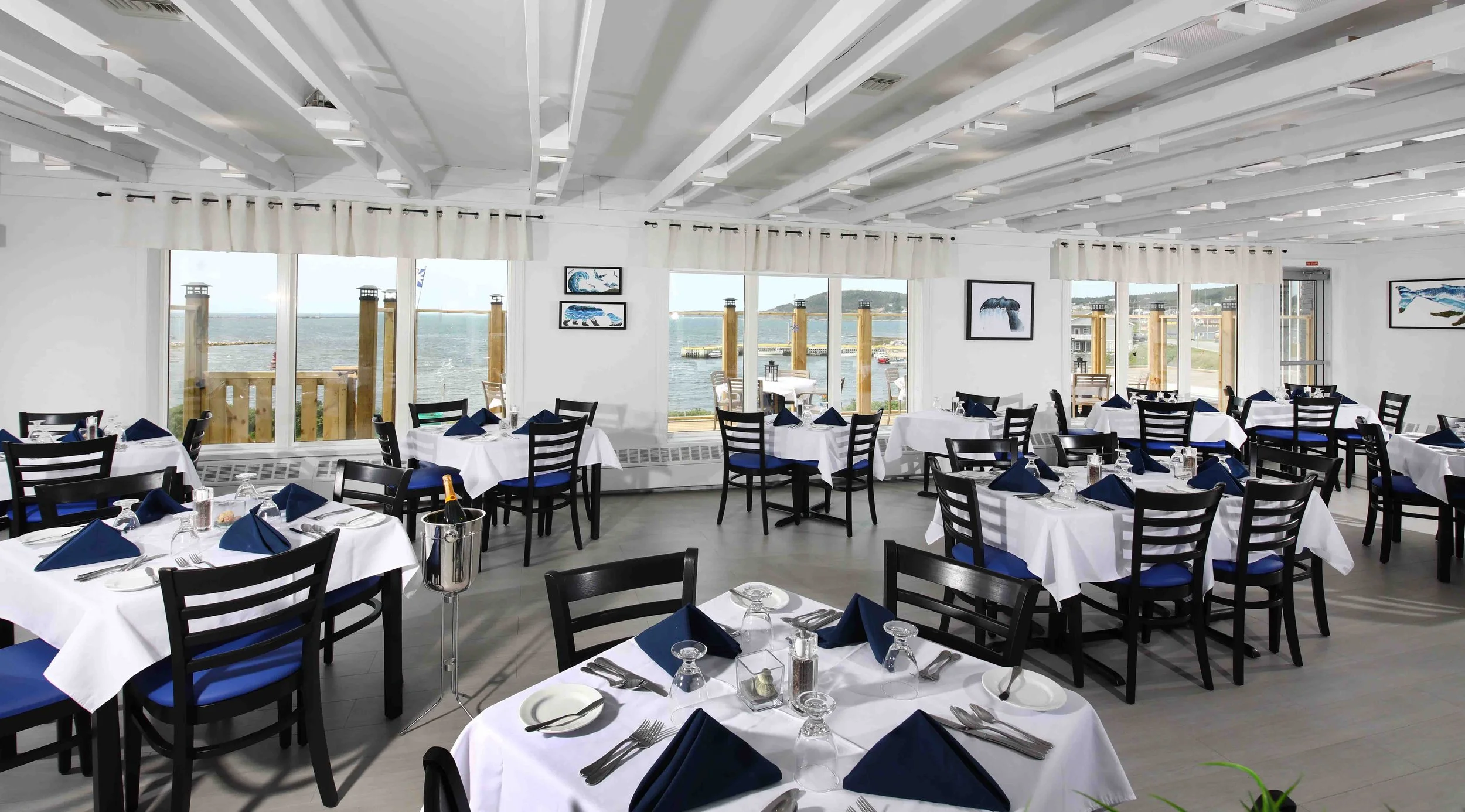 Interior of a seafood restaurant with white walls, large windows overlooking a marina, and tables set with white tablecloths, blue napkins, and glassware.