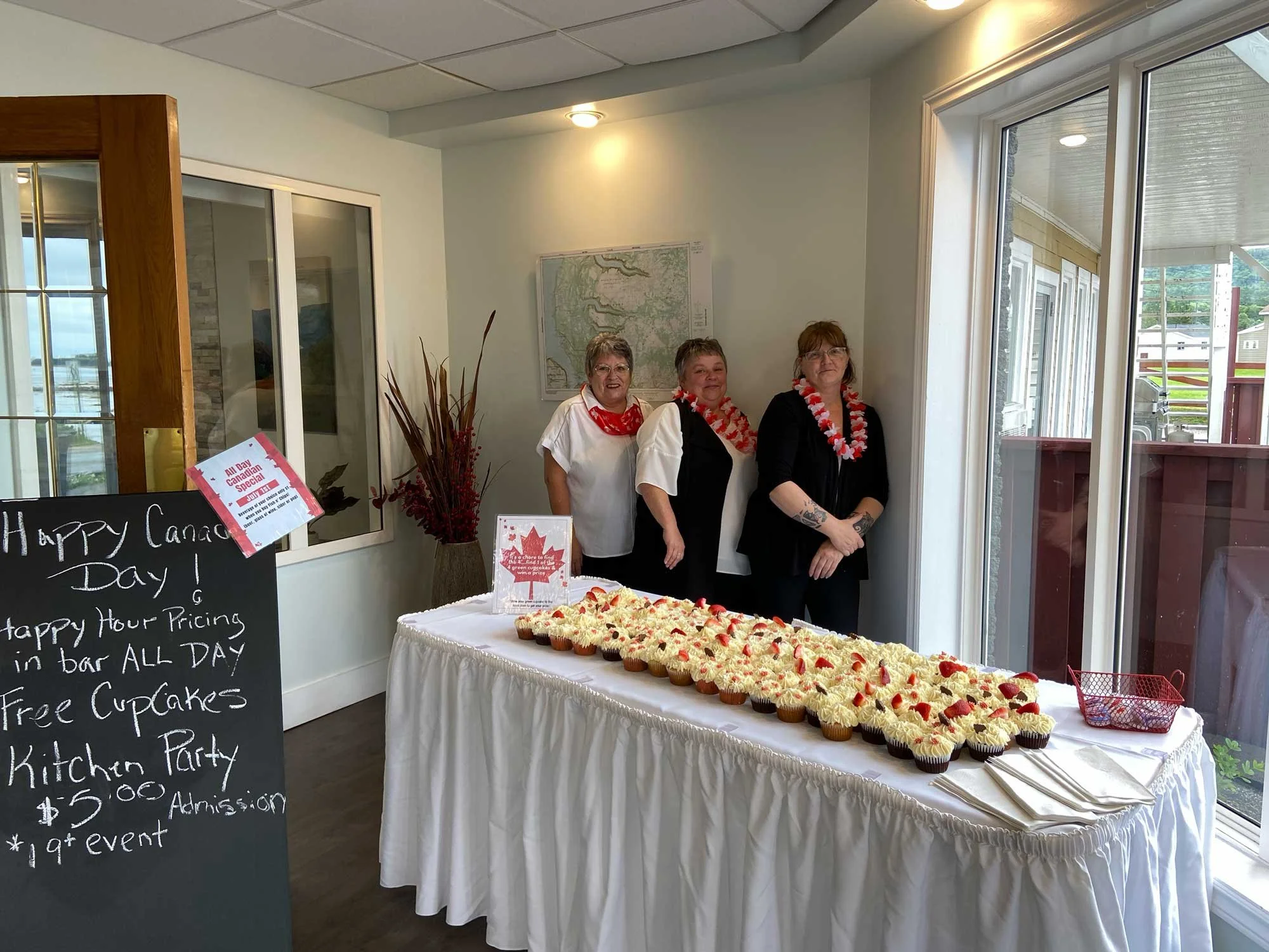 Three women standing behind a table decorated for Canada Day, with numerous cupcakes topped with white frosting and small red decorations. The women are wearing red and white leis, and there is a blackboard sign advertising Canada Day celebrations, i