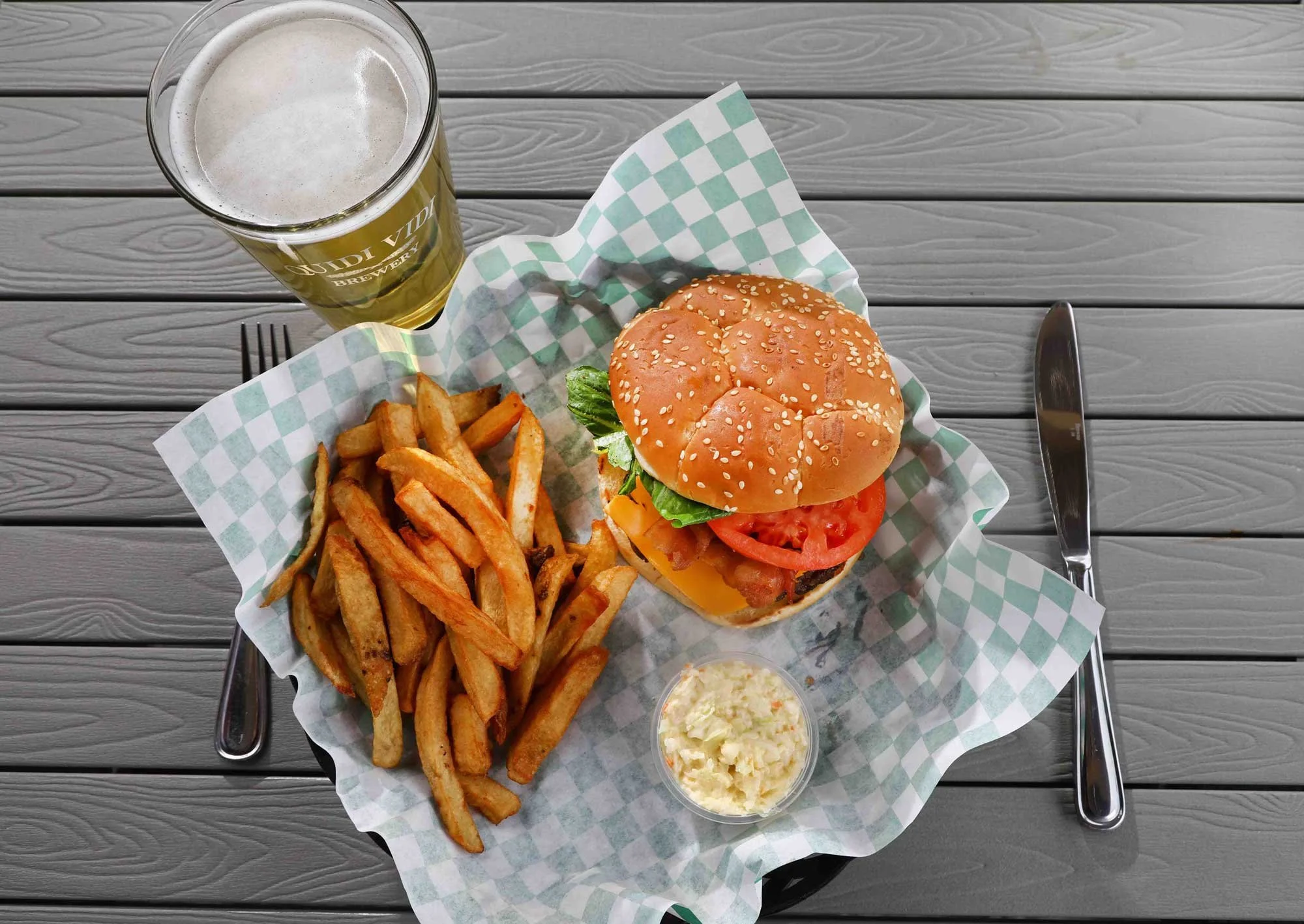 A cheeseburger with lettuce, tomato, and bacon, a side of French fries, a small cup of coleslaw, and a glass of beer on a gray wooden table.