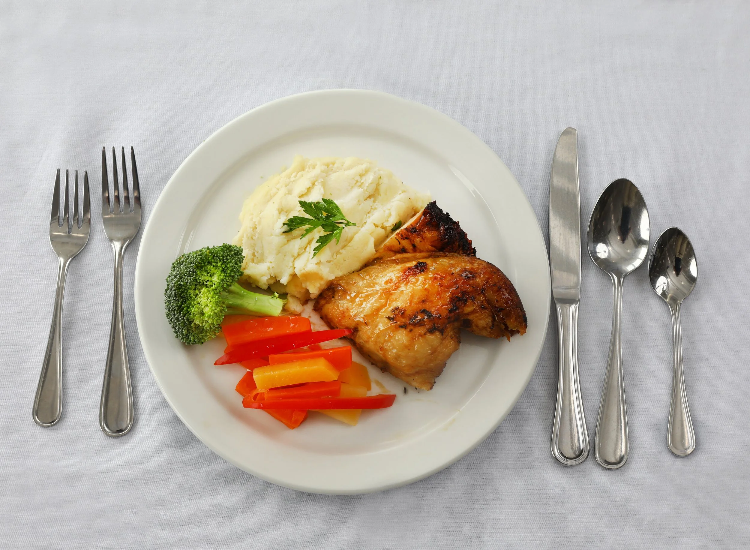 Plate with mashed potatoes, grilled chicken leg, broccoli, and sliced red and yellow bell peppers on a white tablecloth, with forks, knives, and spoons arranged around it.