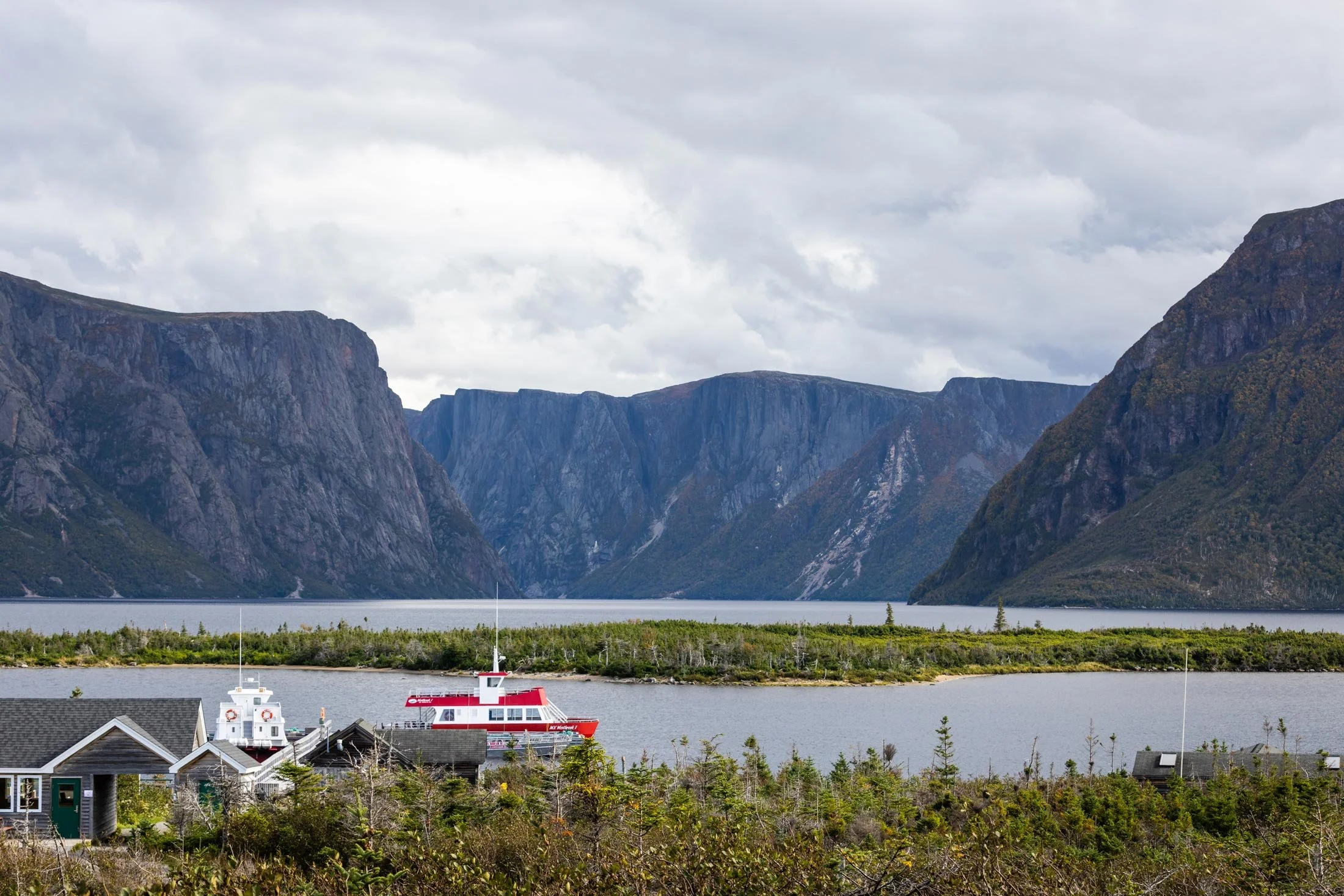 Scenic view of a fjord with mountains in the background, a small boat docked near houses in the foreground, and a cloudy sky overhead.