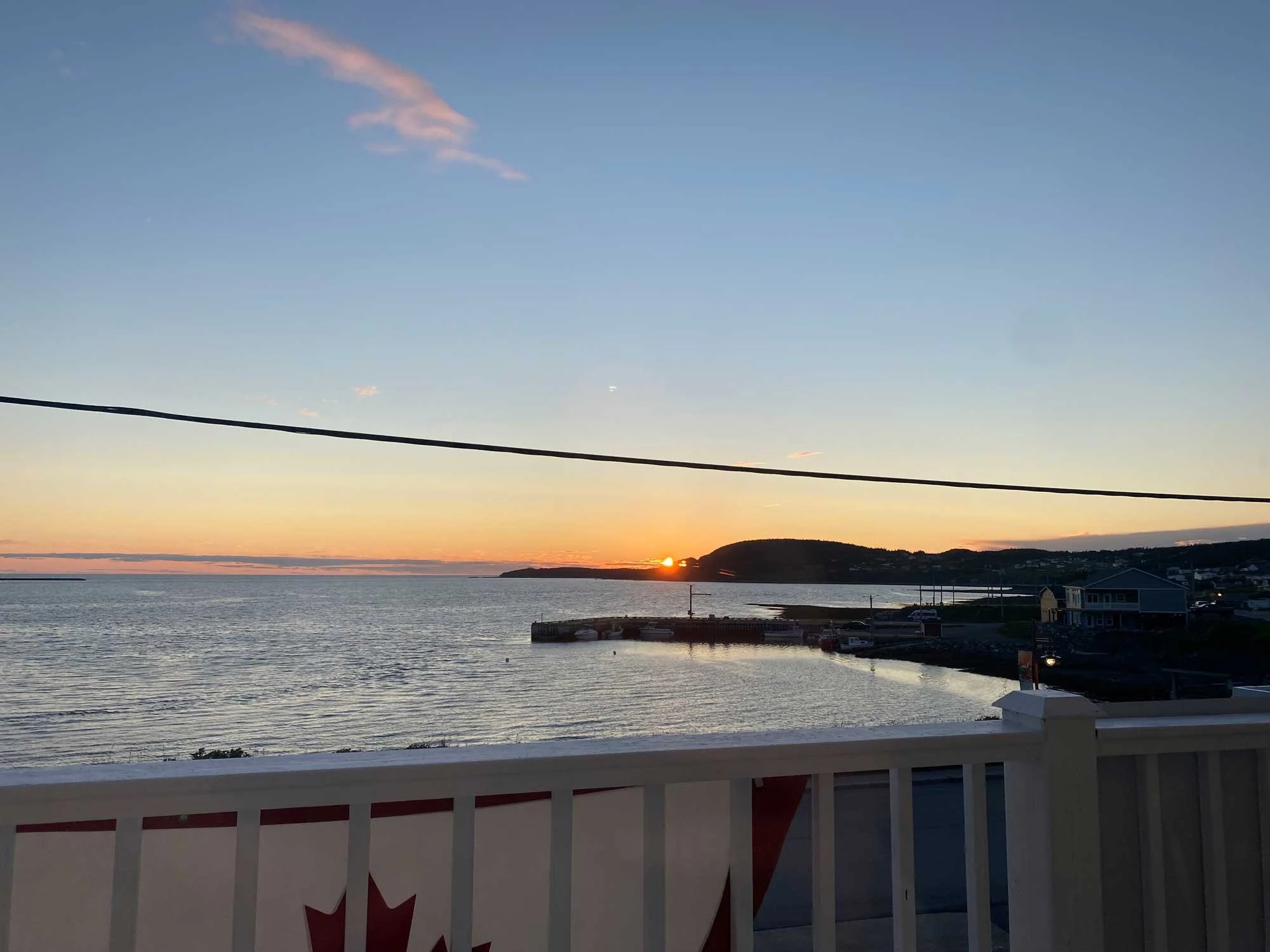 Sunset over water seen from a balcony with a white railing, a Canadian flag, and a distant shoreline with hills and buildings.
