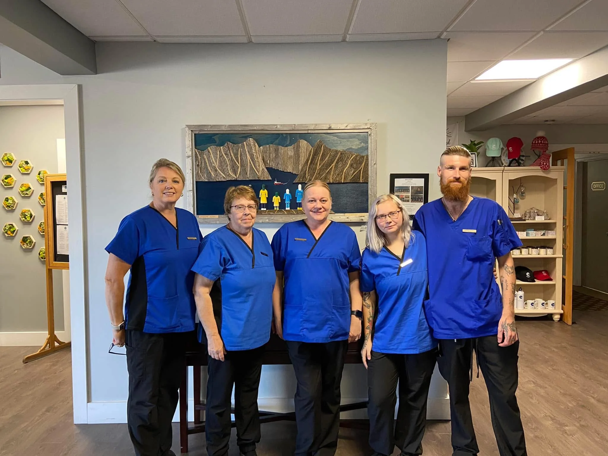 Five staff members wearing blue scrubs standing in a row inside a facility, with artwork on the wall behind them and shelves on the right side.