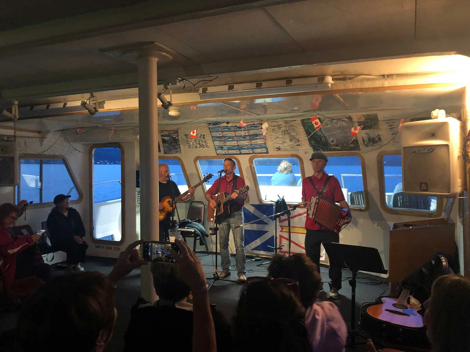 Musicians performing with guitars and an accordion on a boat, with an audience watching, some taking photos. The water is visible through the windows.